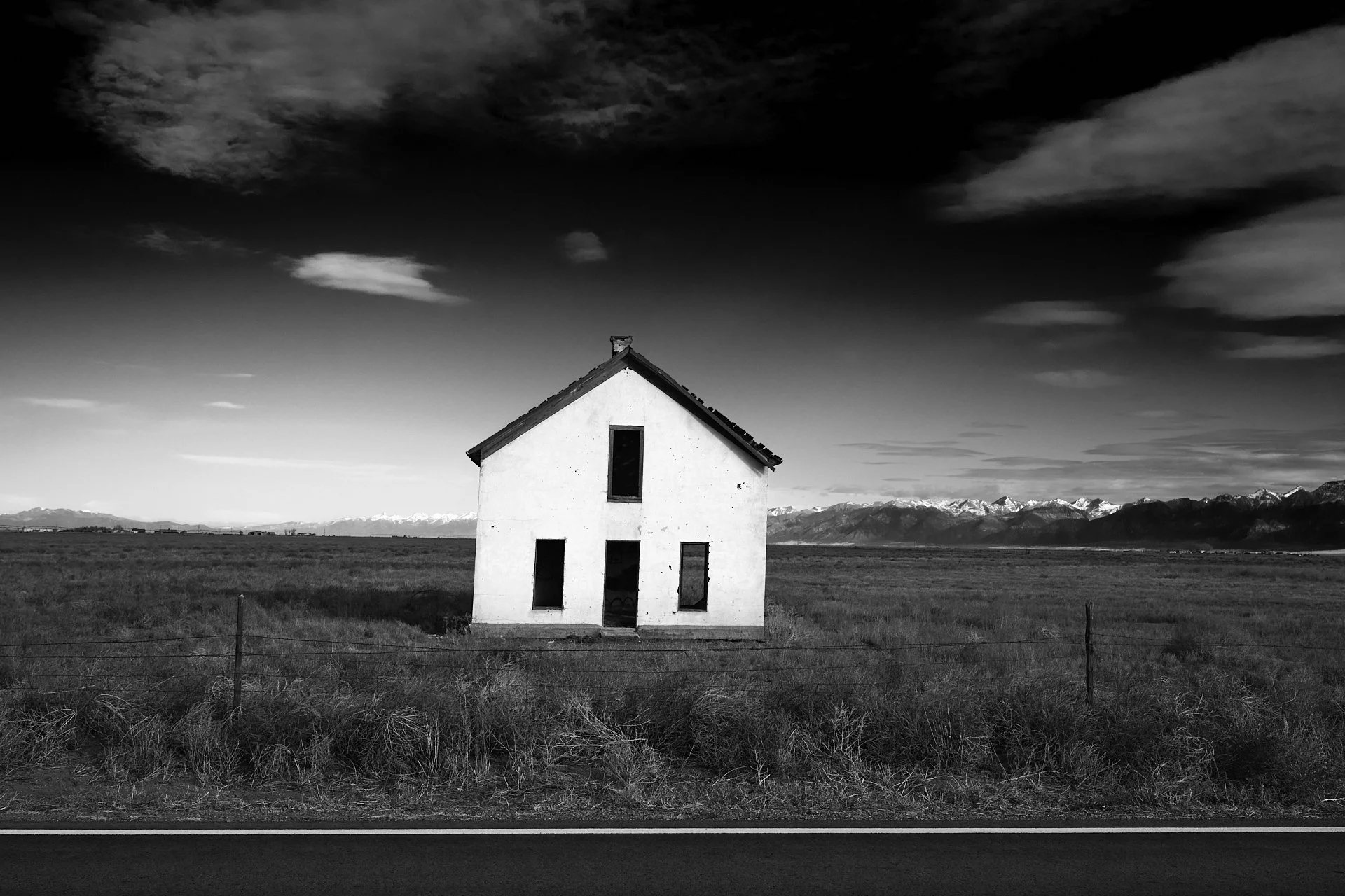 A black and white image of a solitary house with missing windows, standing in a vast open field with mountain range in the background, under a cloudy sky.