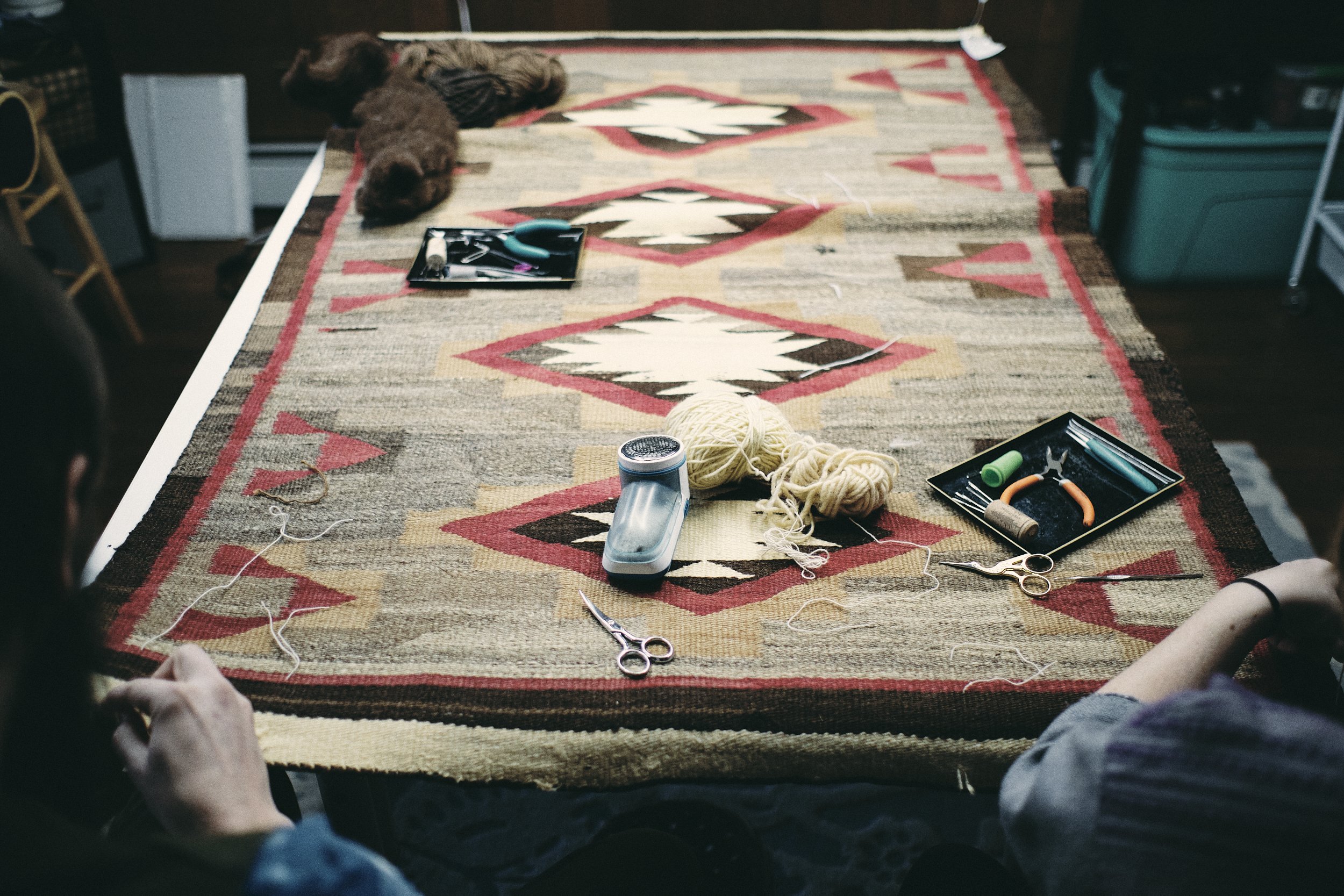 A table covered with a Southwestern patterned rug, holding knitting supplies, scissors, yarn, and tool trays, with two people sitting at the edge of the table.