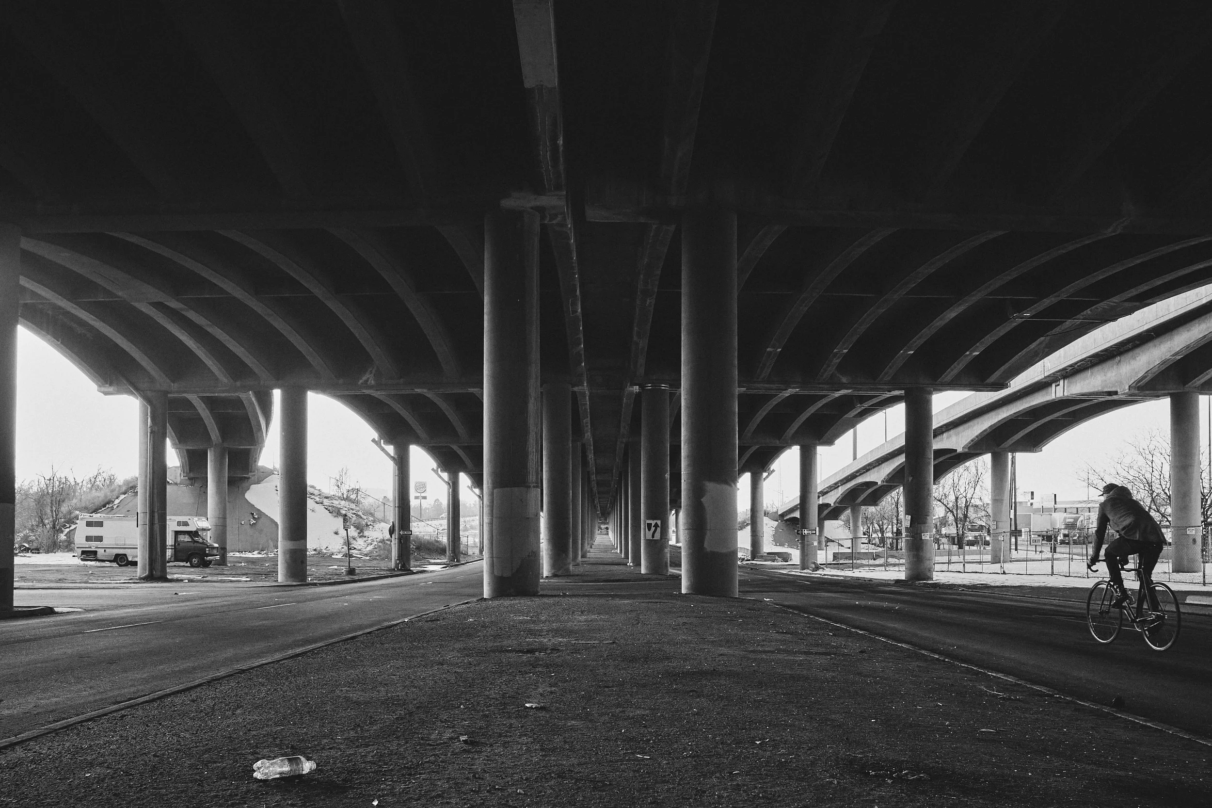 Black and white photo of a cyclist riding under a large bridge with multiple supporting columns, with a van and road in the background and some trees.
