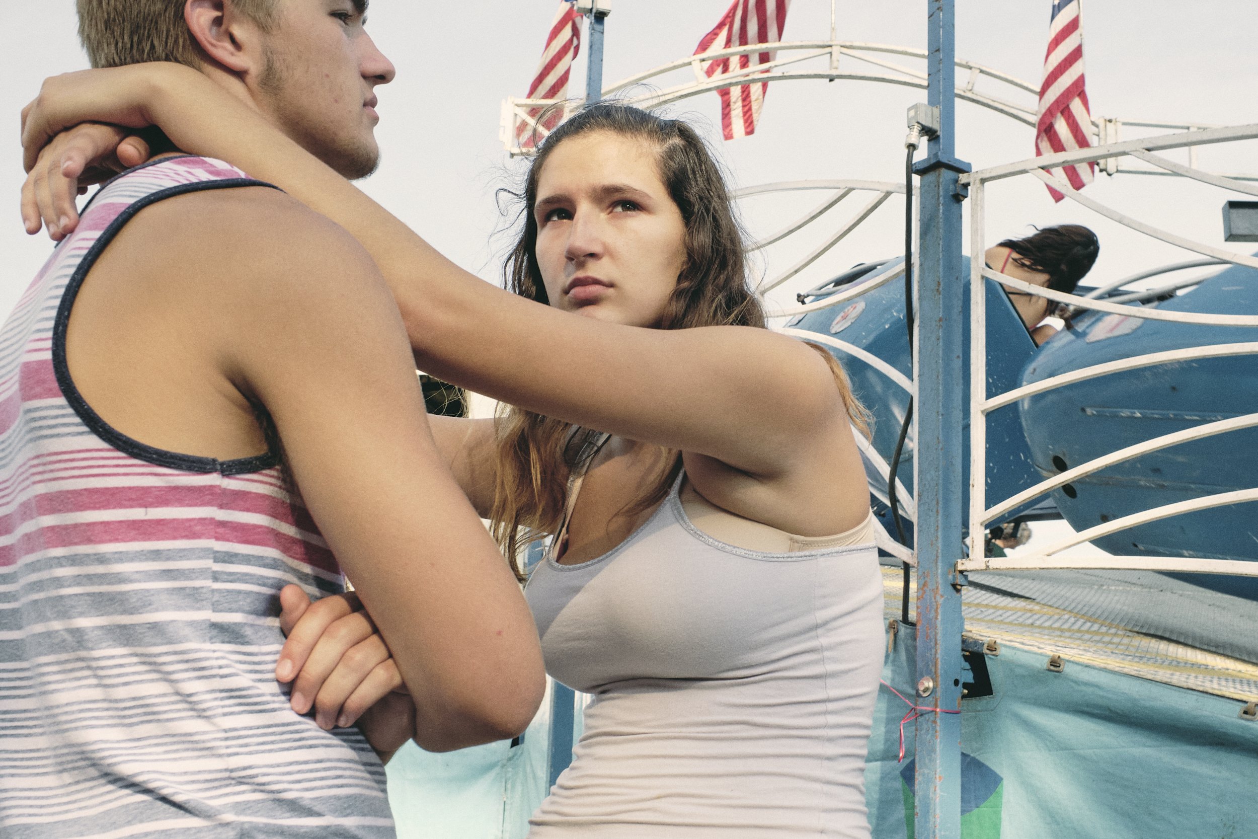 A young woman with brown hair looking upset, arms around a young man in a striped tank top, at a fairground with a blue carnival ride and American flags in the background.