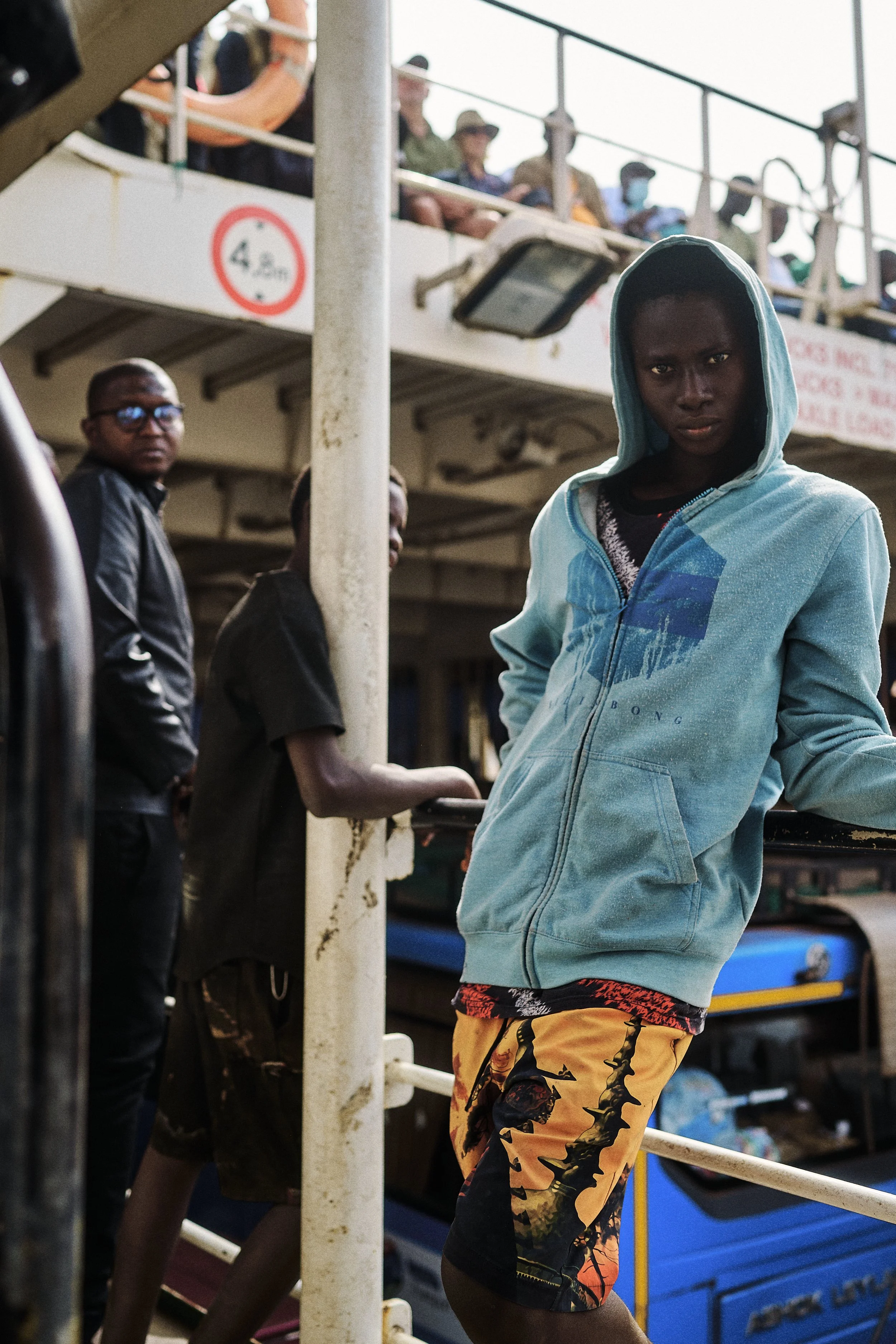 A young person wearing a blue hoodie and colorful shorts leaning against a railing at a busy ferry or boat dock with people waiting in the background.