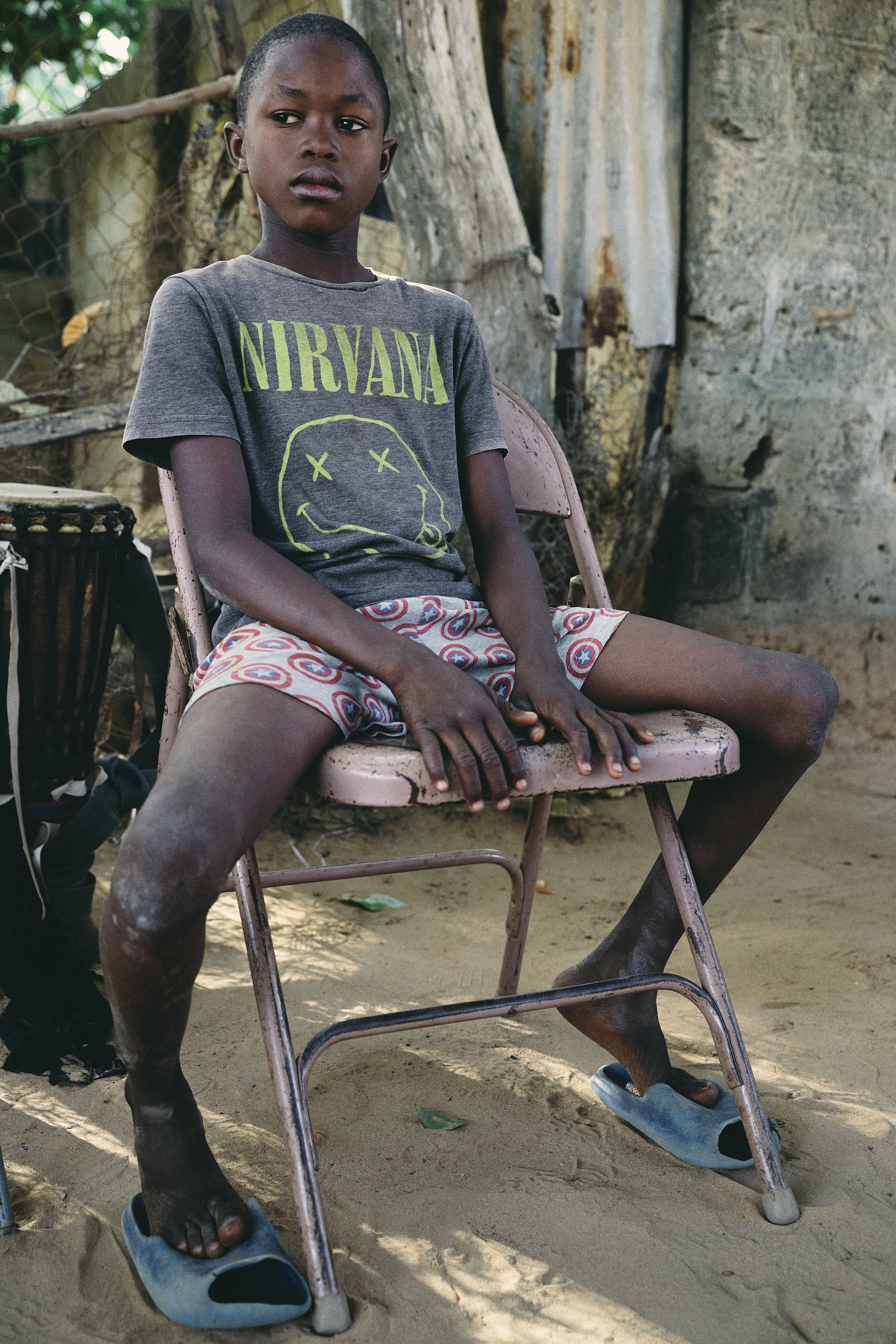 A young boy sitting on a pink metal chair outdoors, wearing a Nirvana T-shirt, patterned shorts, and blue slippers, with a distressed expression on his face.