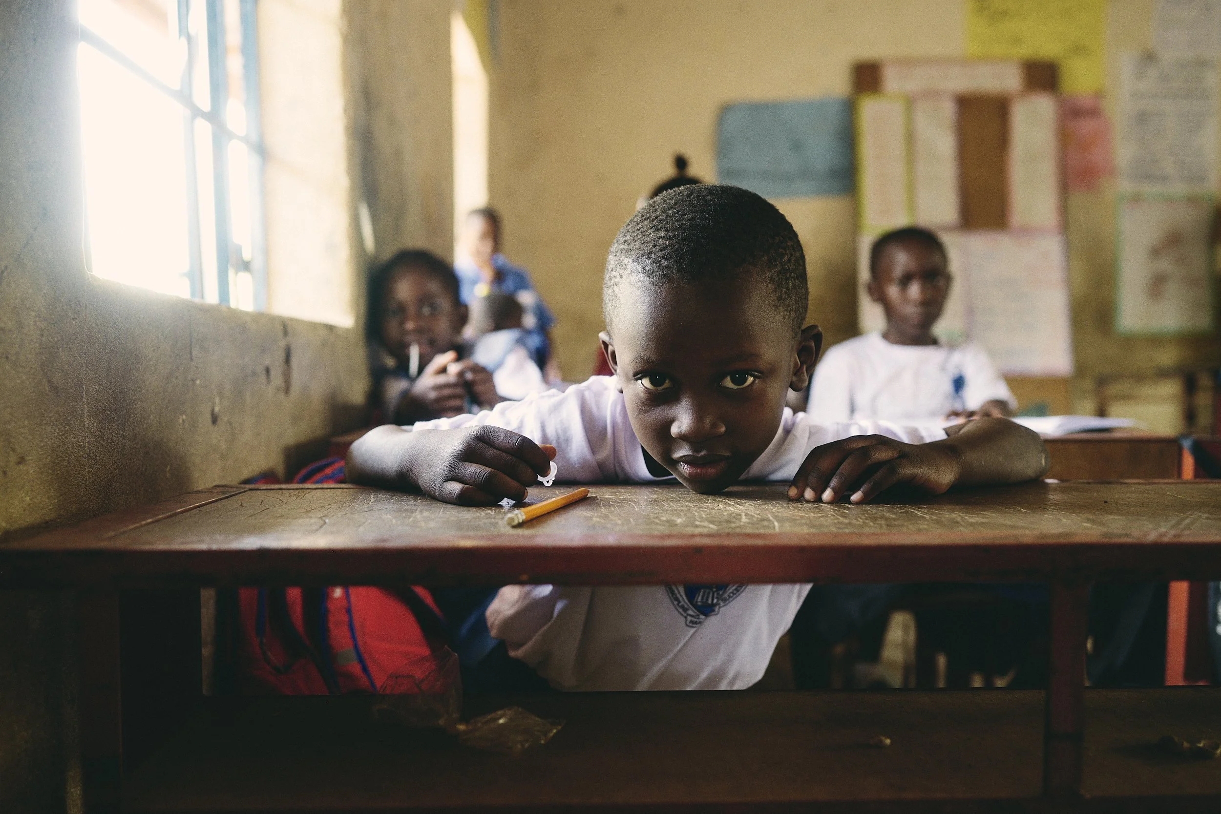 A young boy lying on a desk in a classroom, looking directly at the camera, with other children sitting behind him. The classroom has a window letting in natural light, and educational posters on the wall.