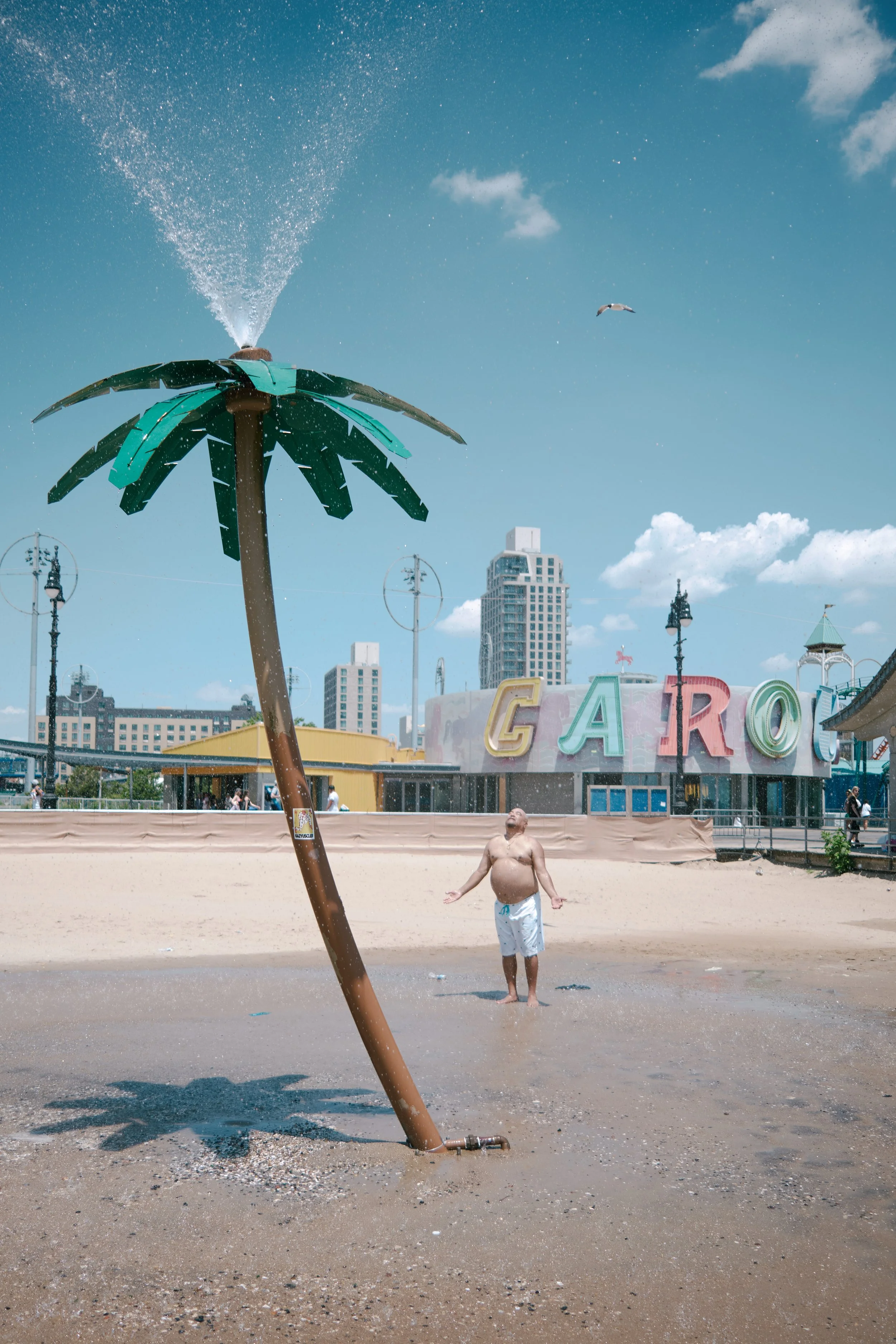 A man standing on a wet sandy beach with a palm tree-shaped sprinkler spraying water, with a colorful amusement park and city skyline in the background, under a partly cloudy sky.