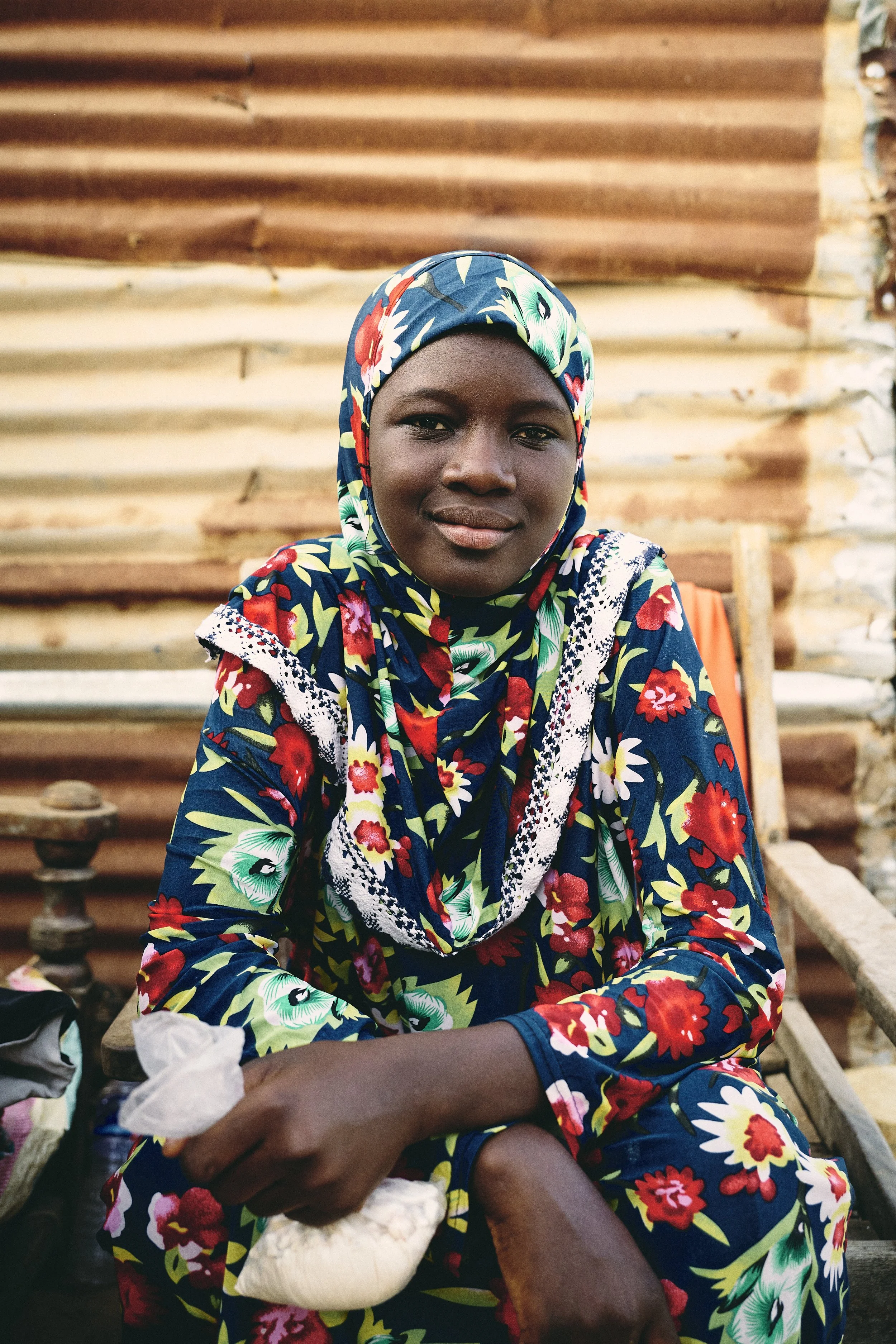 A young woman with dark skin wearing a colorful floral headscarf and matching dress, sitting on a wooden chair outdoors, with a background of corrugated metal sheets.