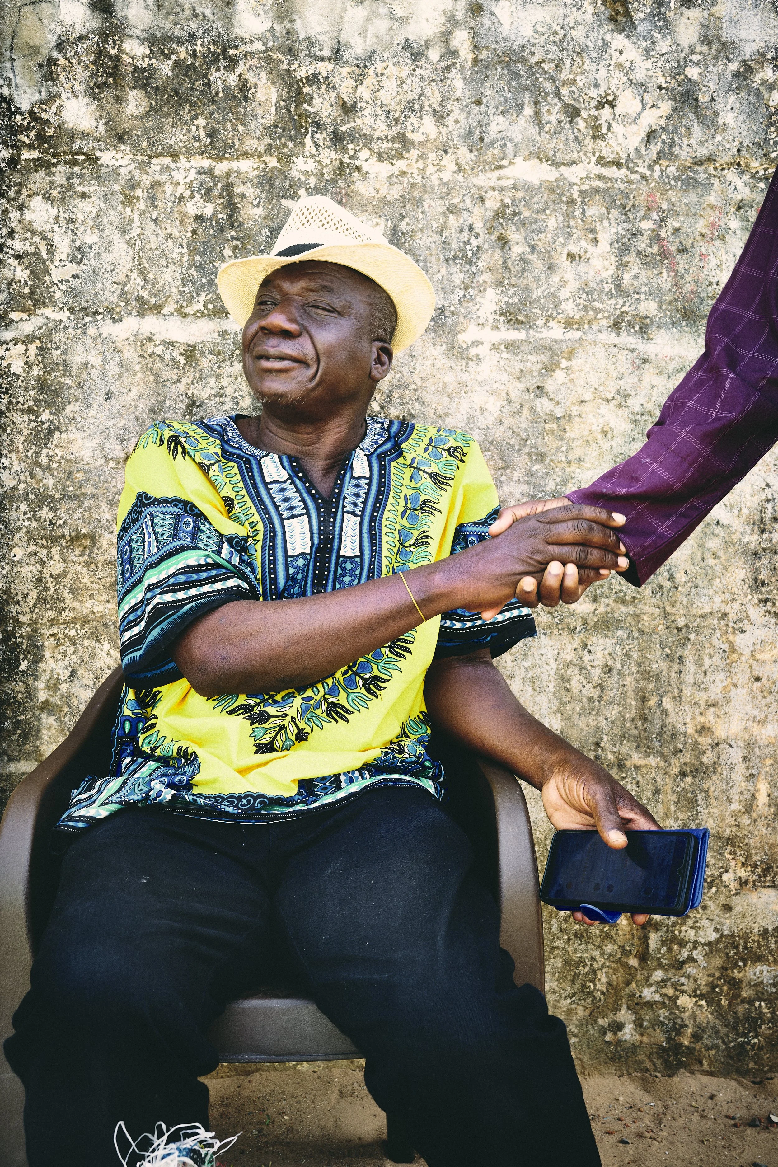 An elderly man wearing a colorful African shirt and a white hat sitting against a textured, weathered wall. He is shaking hands with another person whose arm is extended into the frame, and he is holding a smartphone in his lap.