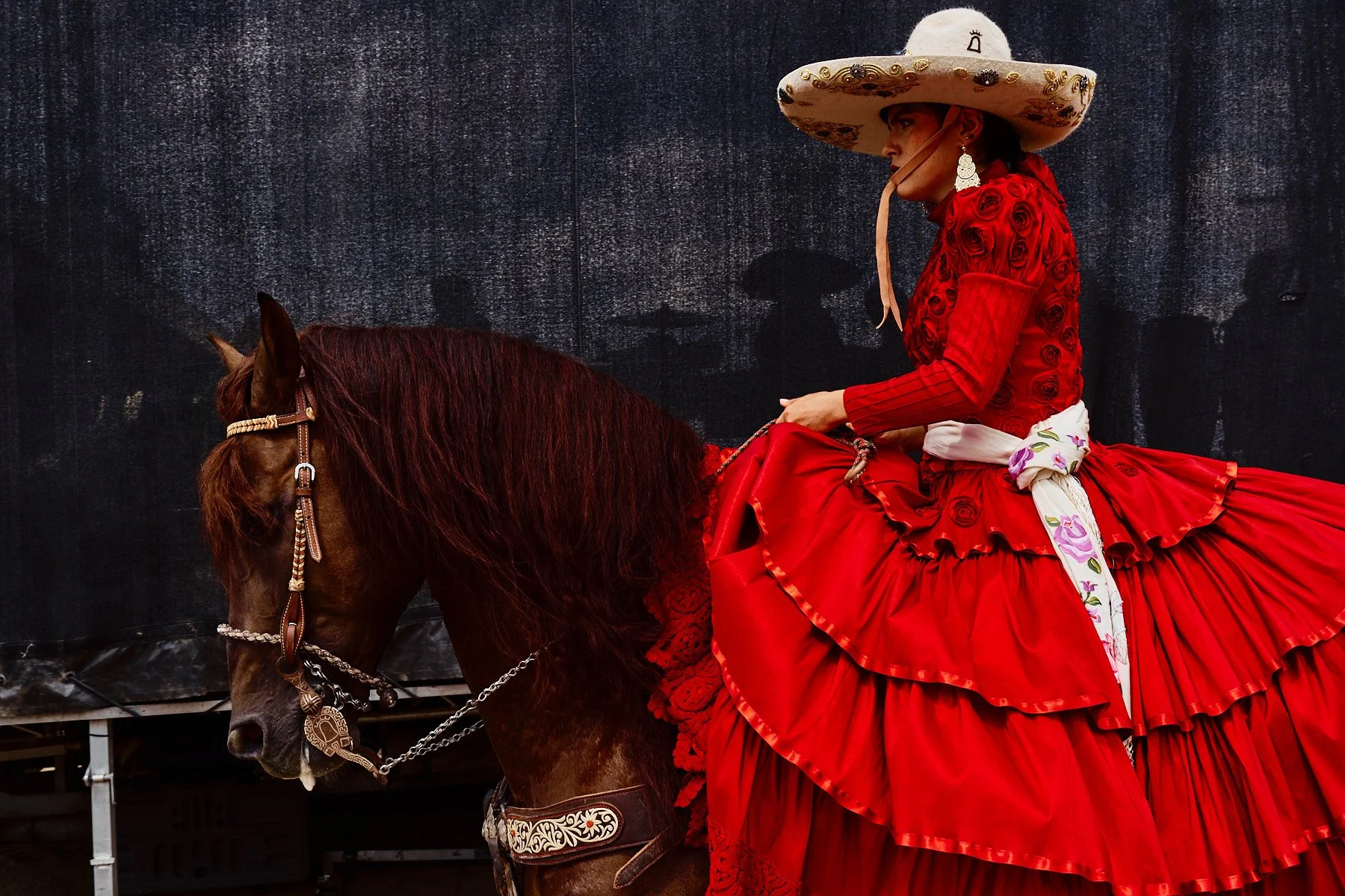A woman dressed in a vibrant red traditional dress and a wide-brimmed sombrero, riding a brown horse with decorative tack, against a dark background.