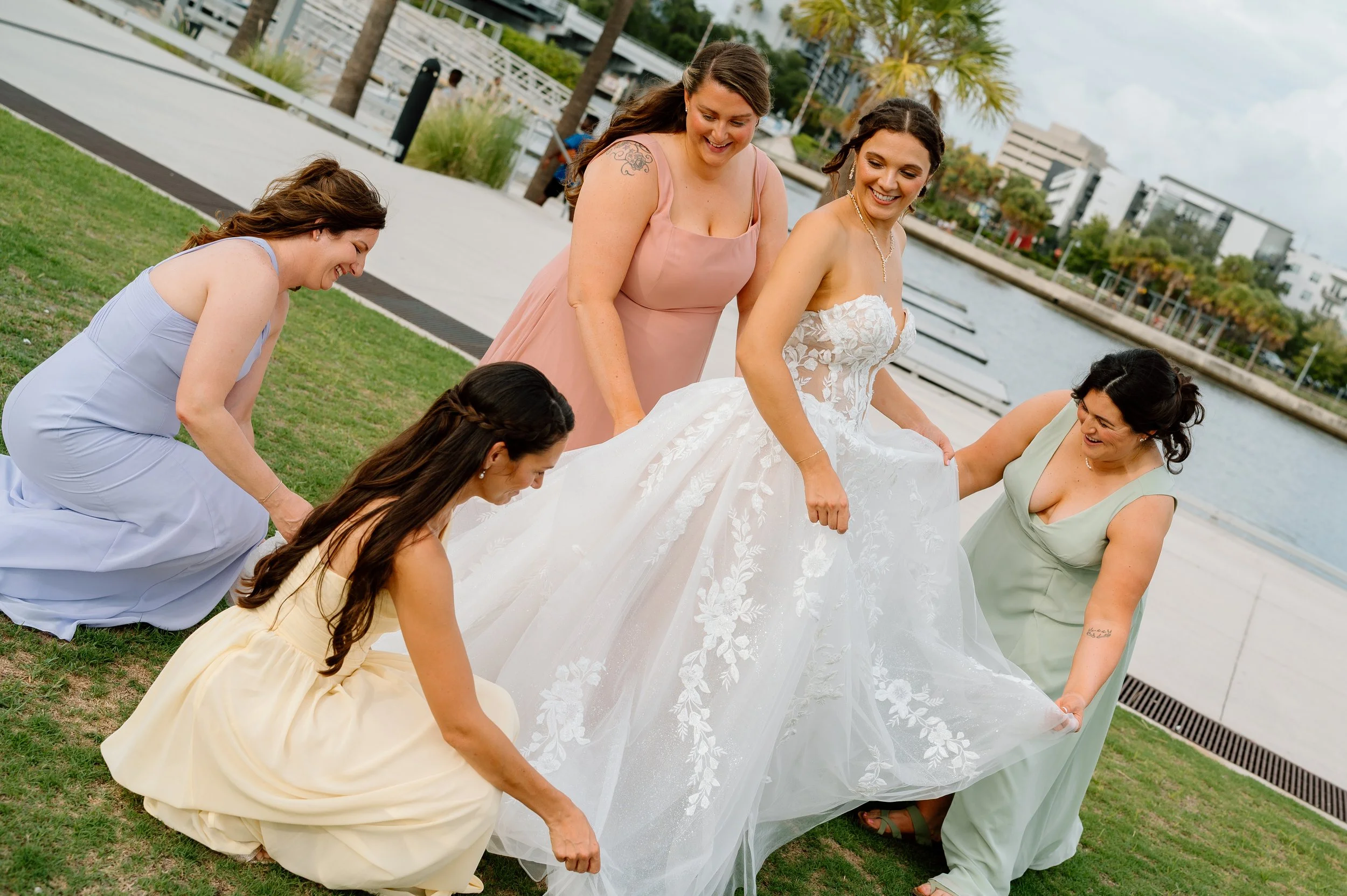 Group of women helping a bride with her wedding dress by the water, with buildings and trees in the background. The group is on the Tampa Riverwalk at the Buckhorn Tampa Rivercenter for a wedding.