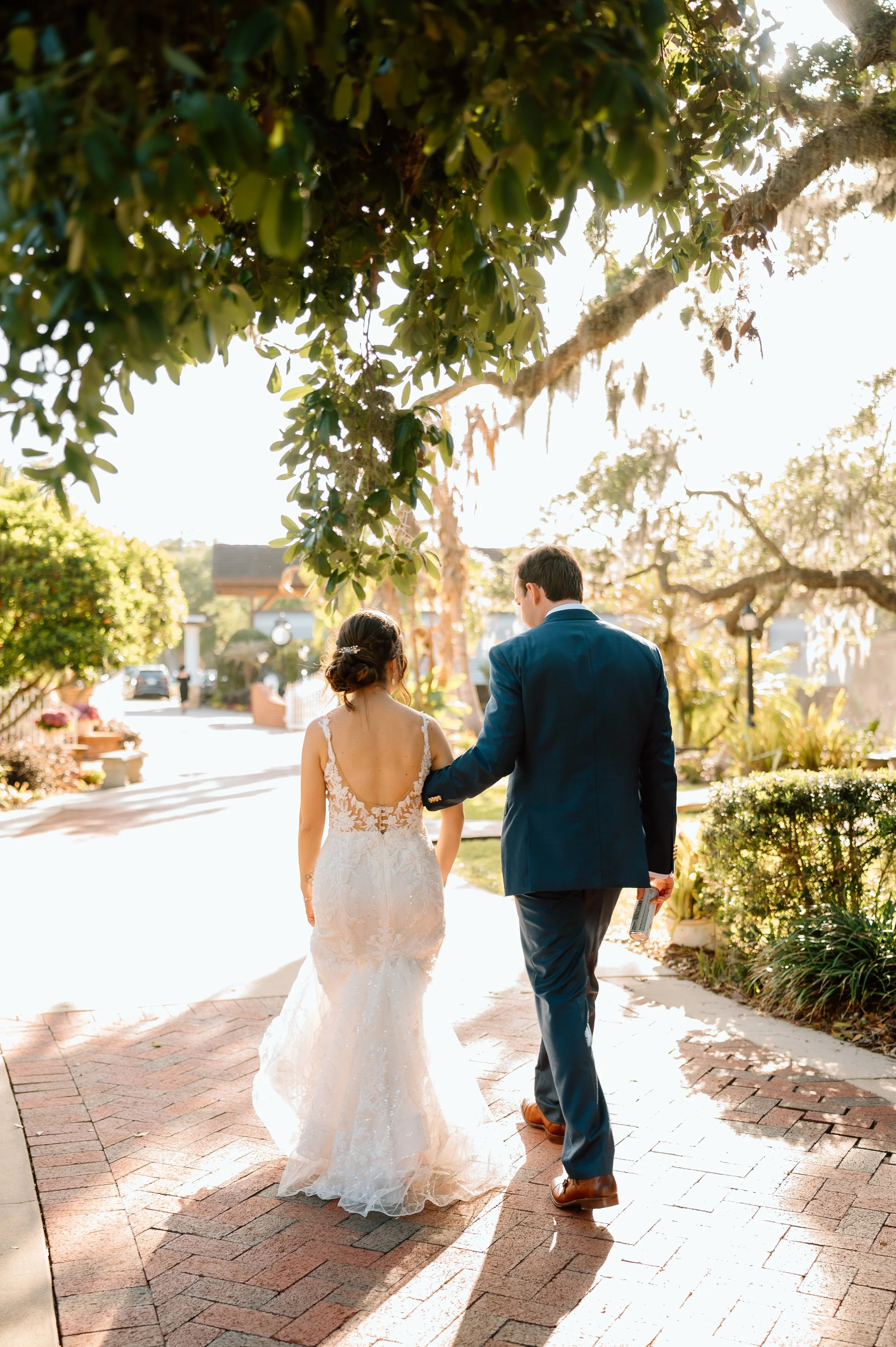A bride and groom walking together down a sunlit brick pathway in a garden, holding hands with the groom holding a microphone in his right hand. The bride wears a white lace wedding gown with a low back, and the groom is dressed in a blue suit. They 