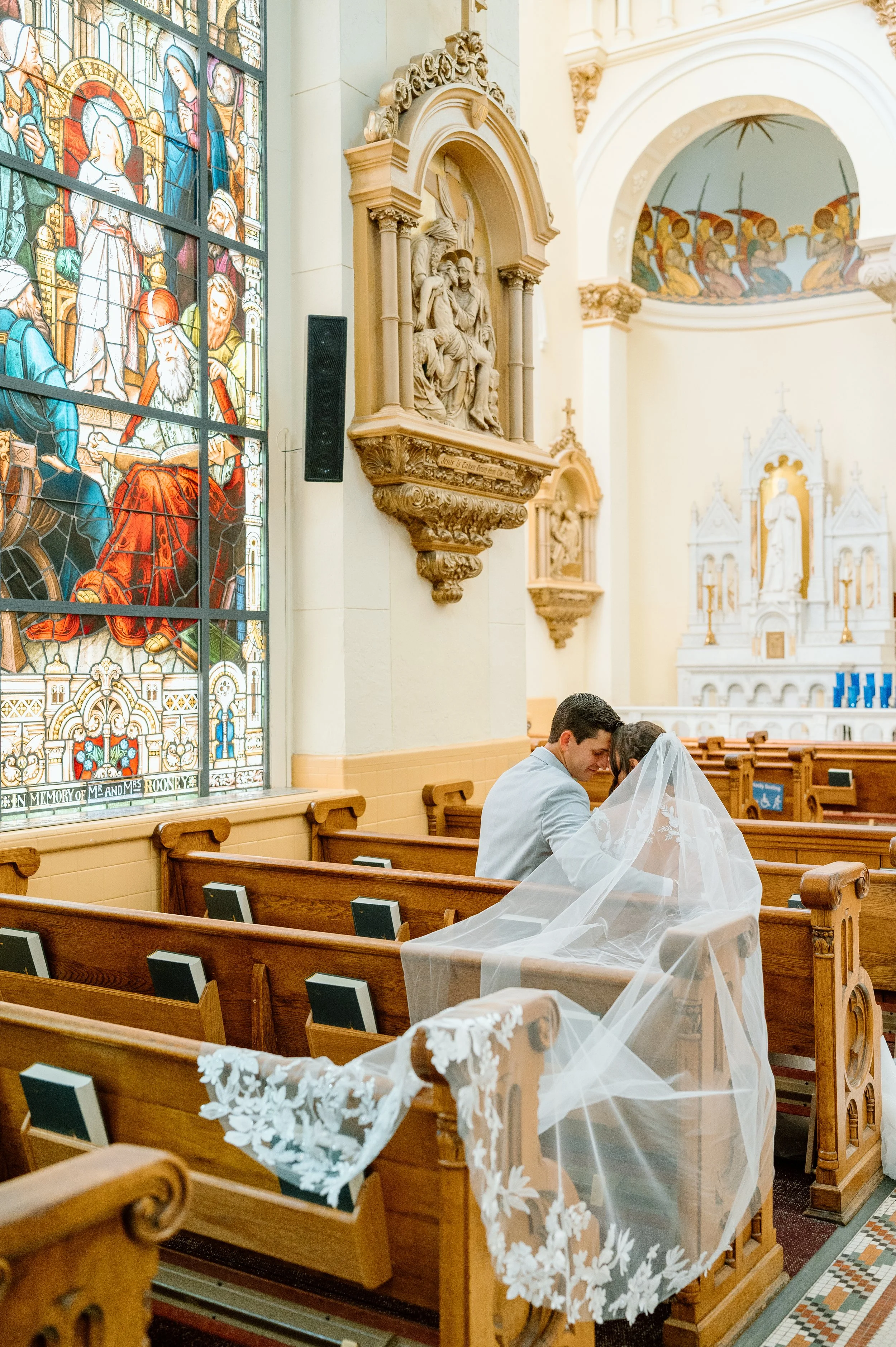 A Tampa, Florida church interior with stained glass windows depicting religious figures, a statue of Jesus with a woman at his side, and a bride and groom sitting in wooden pews, with the bride wearing a veil that drapes over the pew.