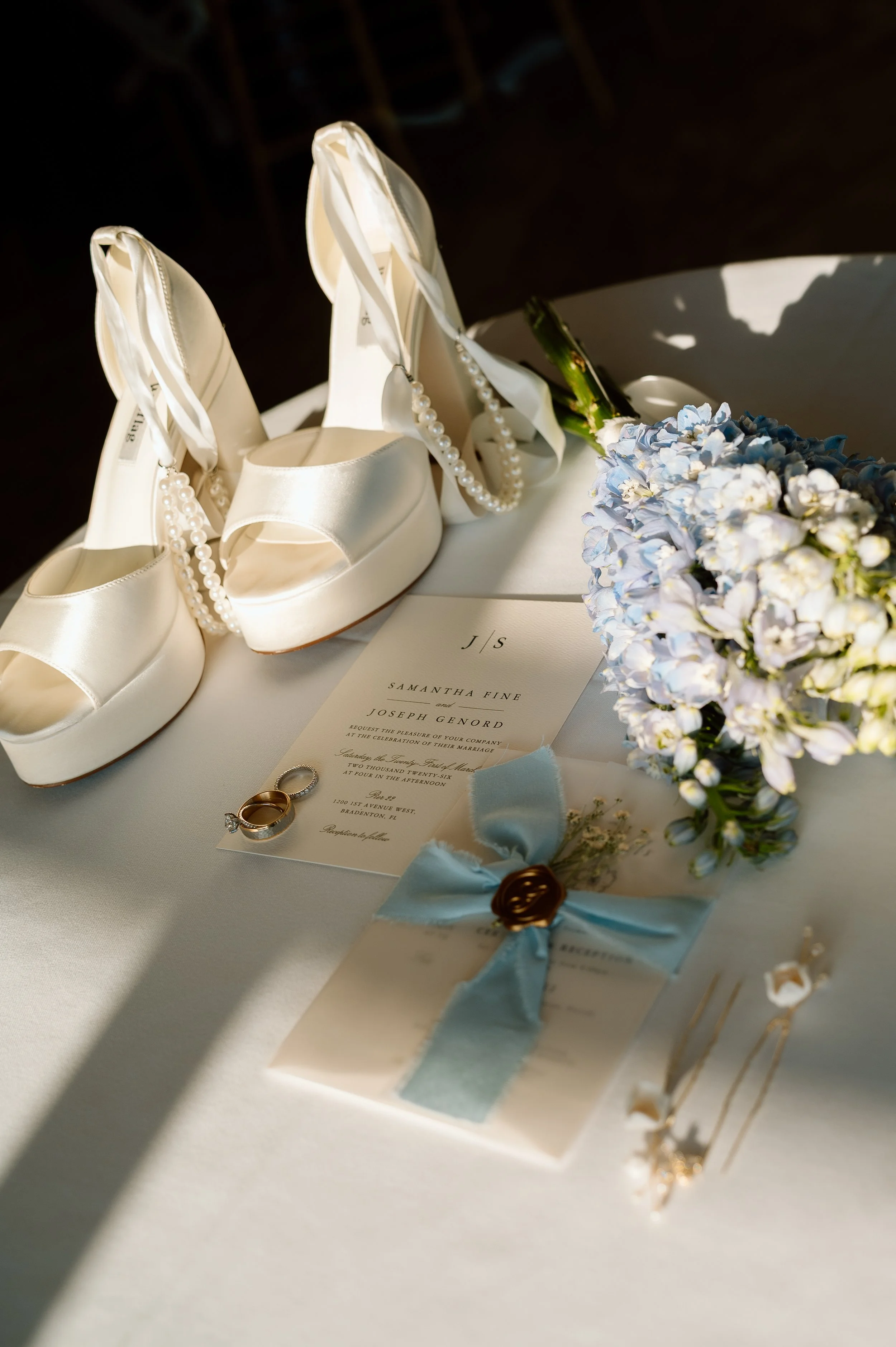 White high-heeled shoes with pearl anklets, wedding rings, a bridal bouquet of blue and white flowers, and wedding invitations on a white surface.