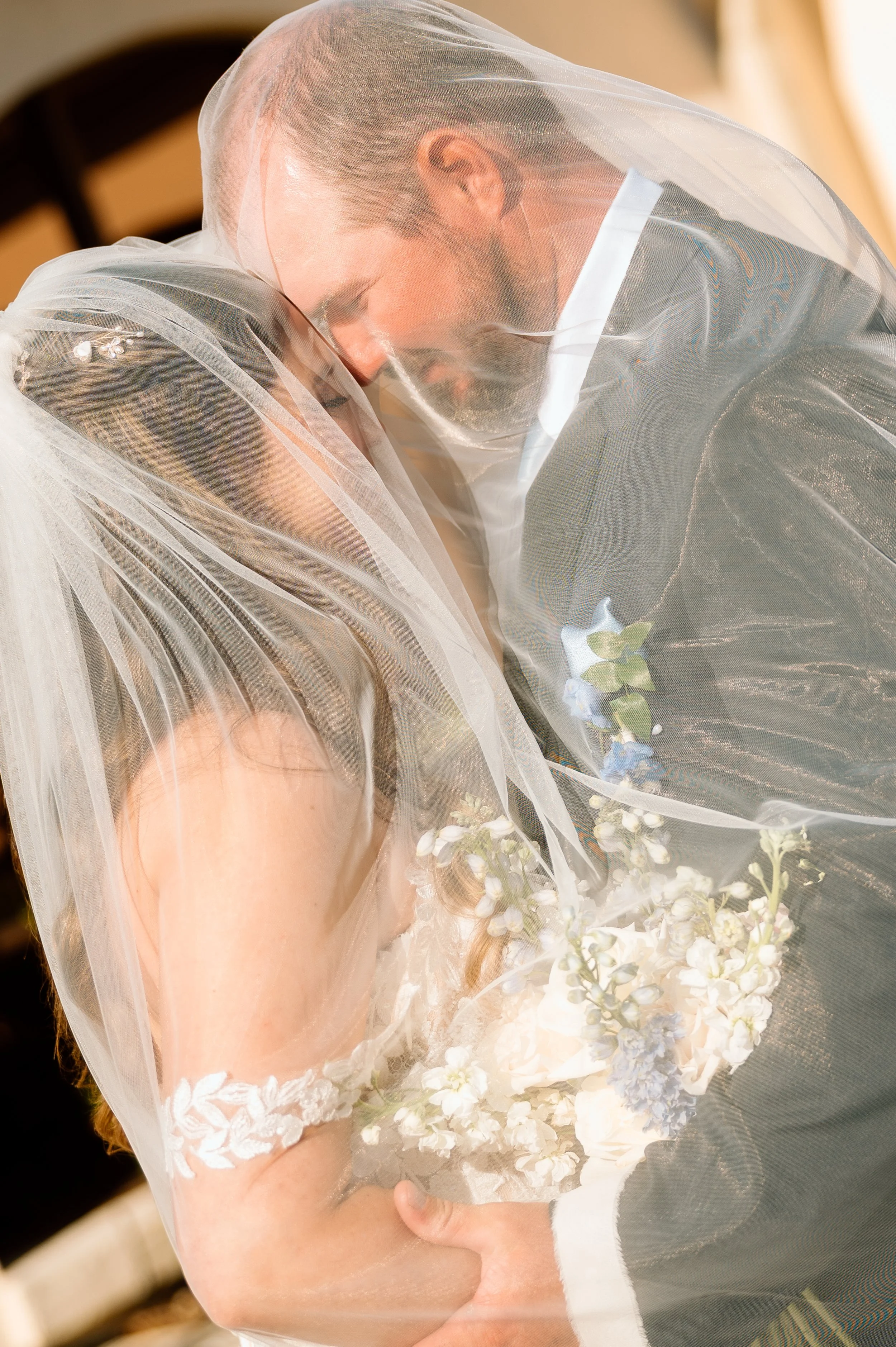 A bride and groom Share a tender embrace with their foreheads touching, both wearing wedding attire, with the bride holding a bouquet of flowers and a sheer veil covering both of their heads.