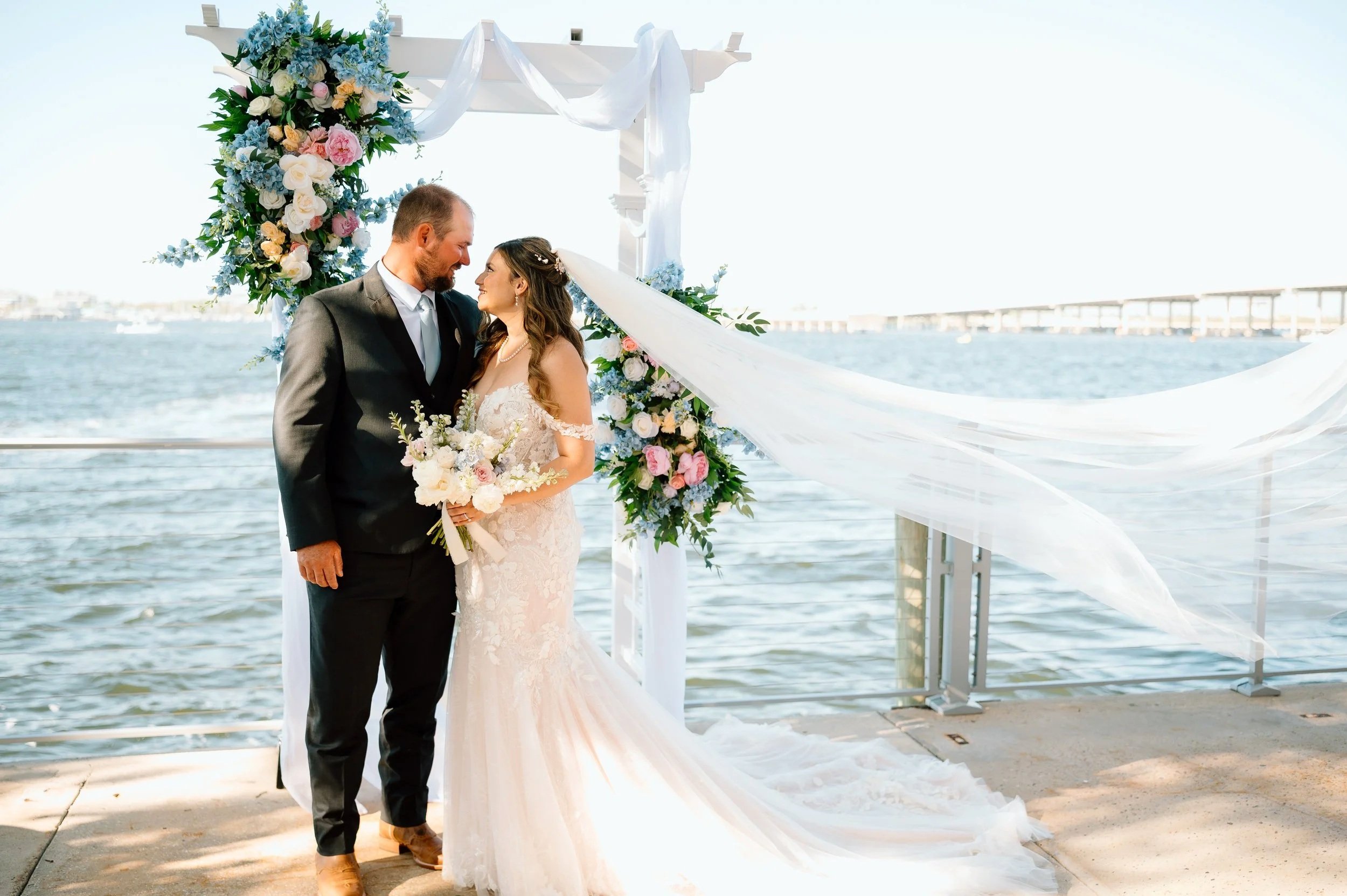 A bride and groom standing close together, smiling, on a wedding altar at Pier 22 in Bradenton, Florida. The venue is decorated with flowers near a body of water with a bridge in the background.