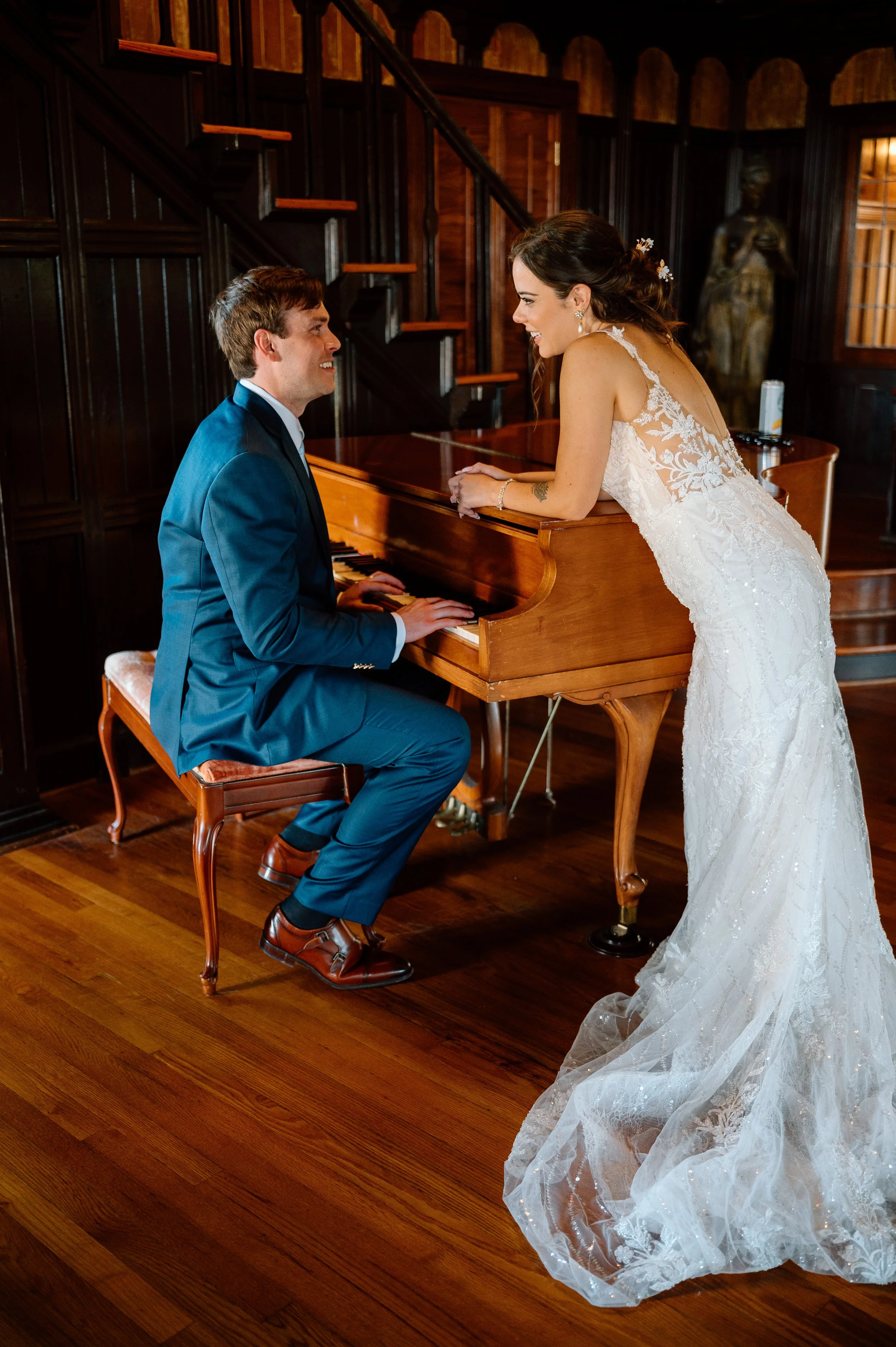 A bride in a white lace wedding gown leaning over a piano, smiling at a groom in a blue suit who is seated and playing the piano, inside a wooden room with dark-paneled walls.