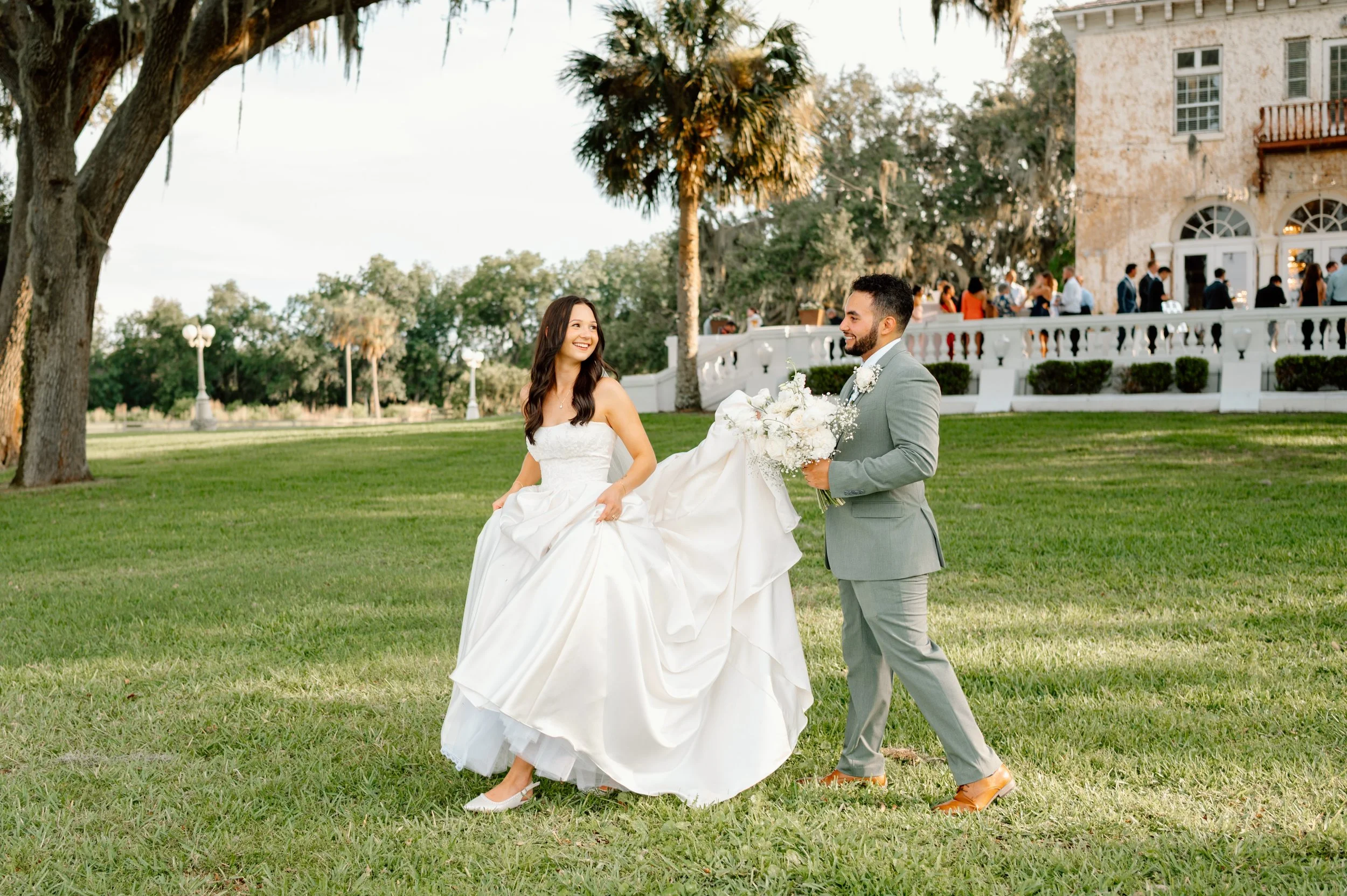 A bride in a white wedding dress and a groom in a light gray suit exchanging wedding vows outdoors. There are guests and a historic building in the background, surrounded by trees and a lush green lawn at Bella Cosa wedding venue in Lake Wales, FL.