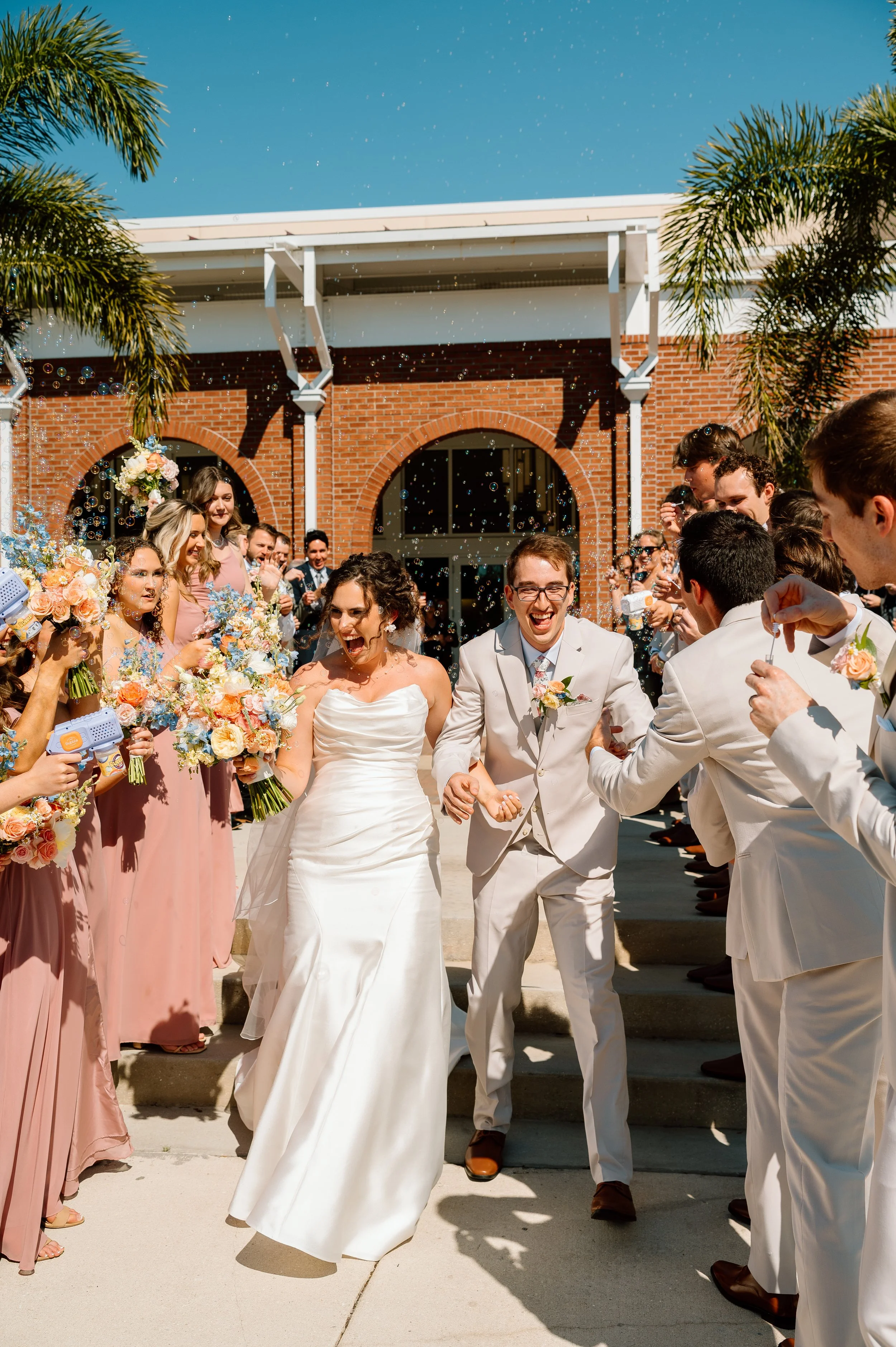 A wedding celebration outdoors with a bride and groom walking down stairs, surrounded by guests, with bubbles floating in the air, and a brick building with arched windows in the background.