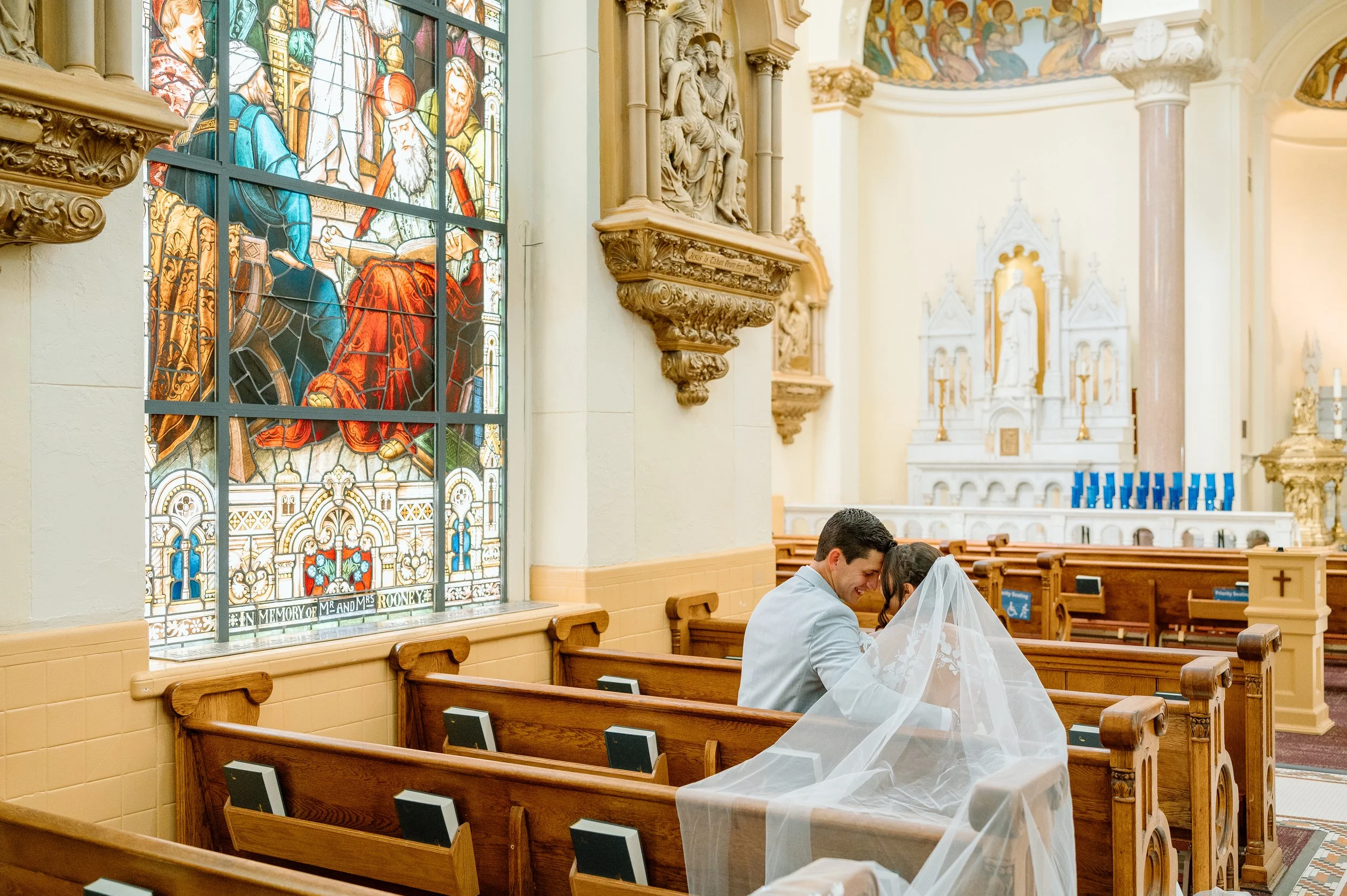 A newlywed couple sitting in a church, smiling at each other. The Sacred Heart Church in Tampa, FL has a stained glass window and ornate decorations.