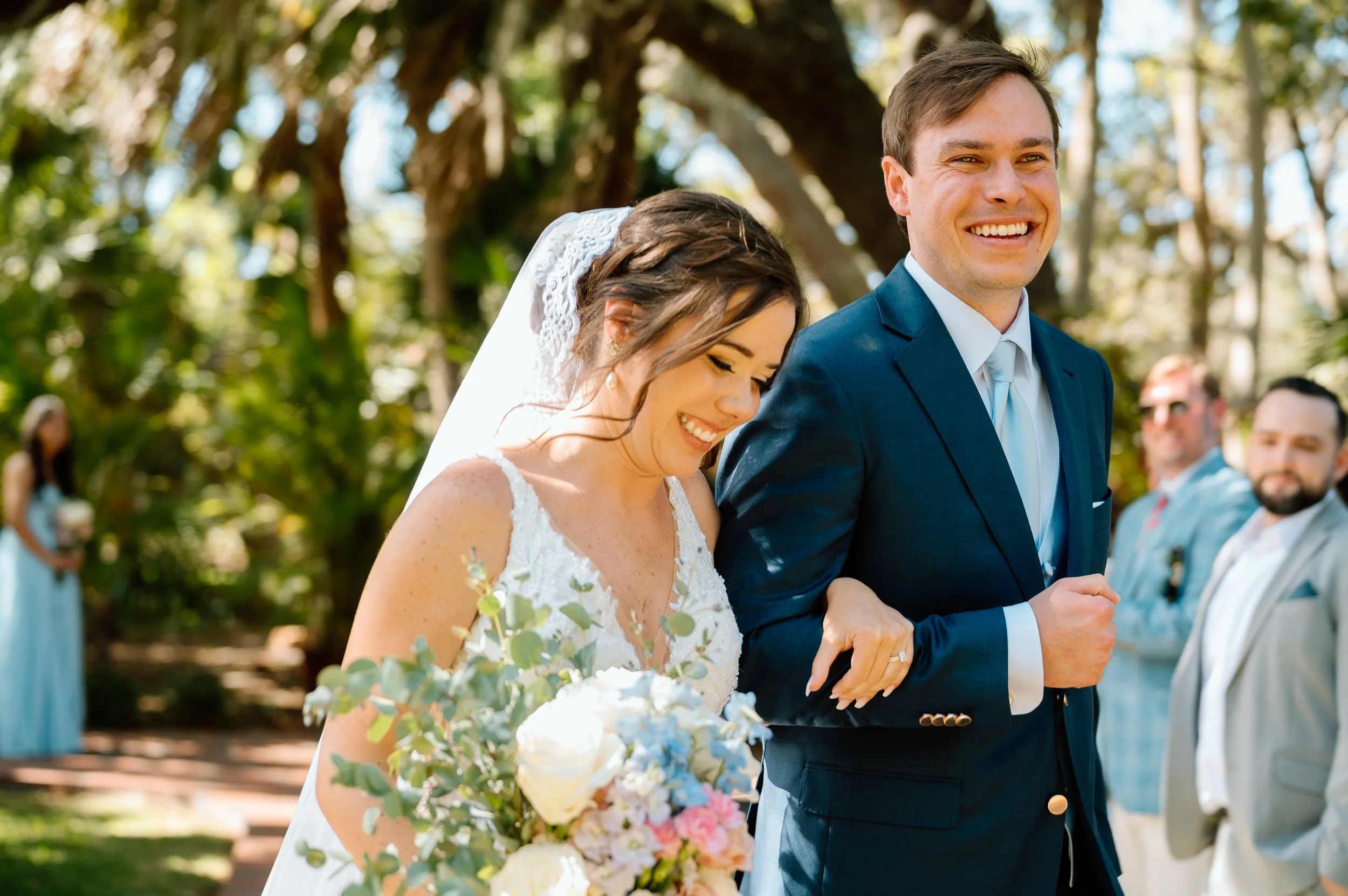 A bride and groom smiling and holding hands during an outdoor wedding ceremony surrounded by trees and guests.