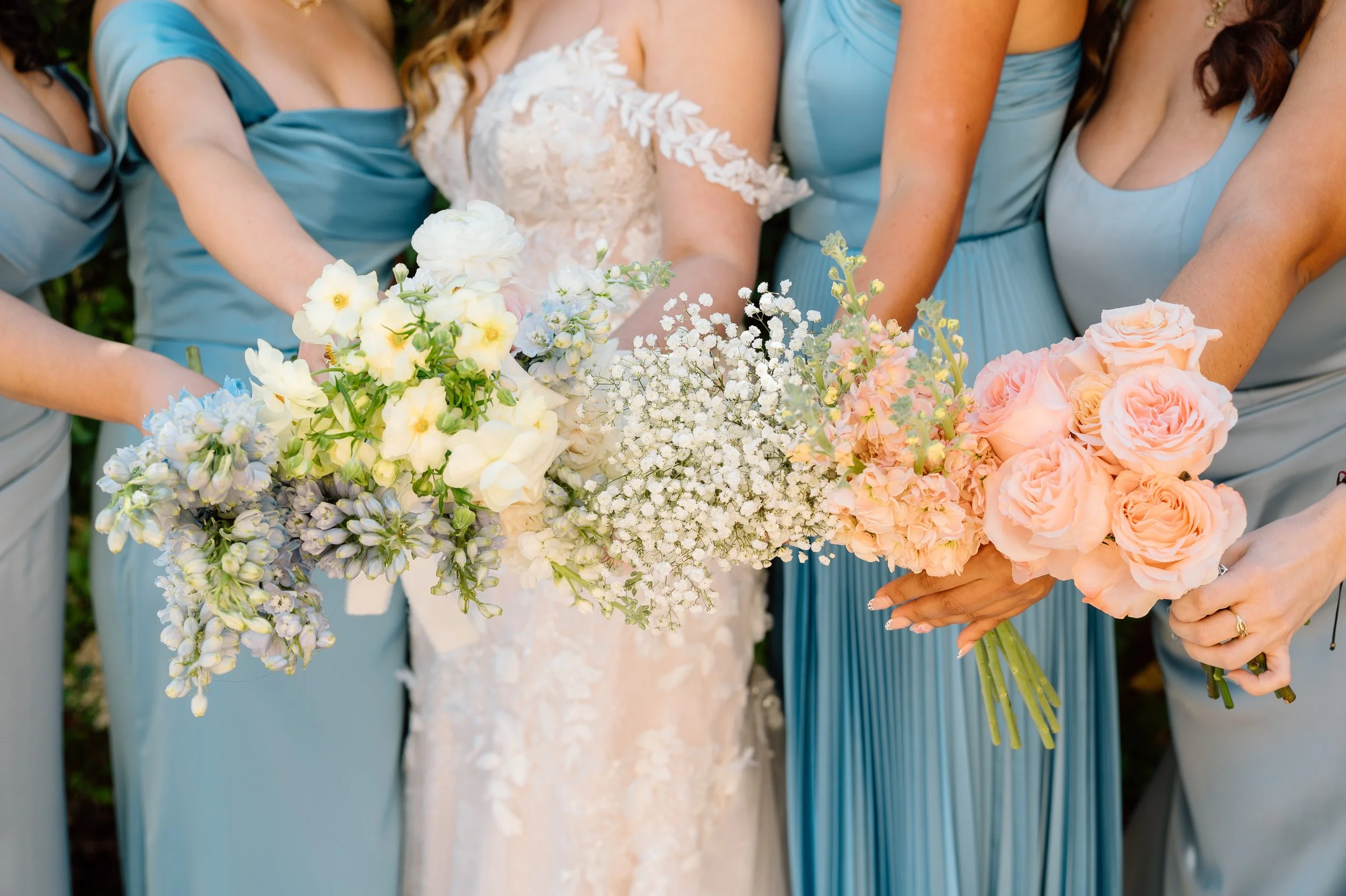 Close-up of women holding bouquets of white and peach flowers at a wedding, with one woman in a white lace wedding dress and others in blue dresses.