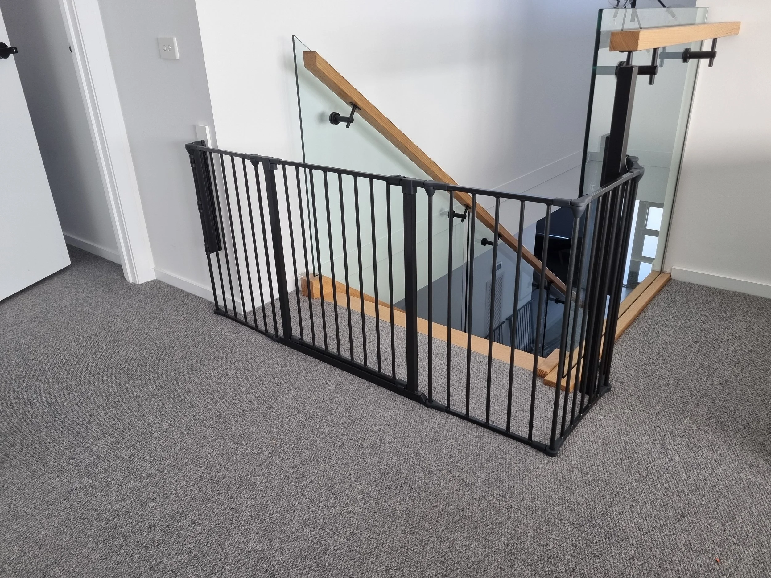 Black L-shaped baby gate at the top of stairs with a glass balustrade