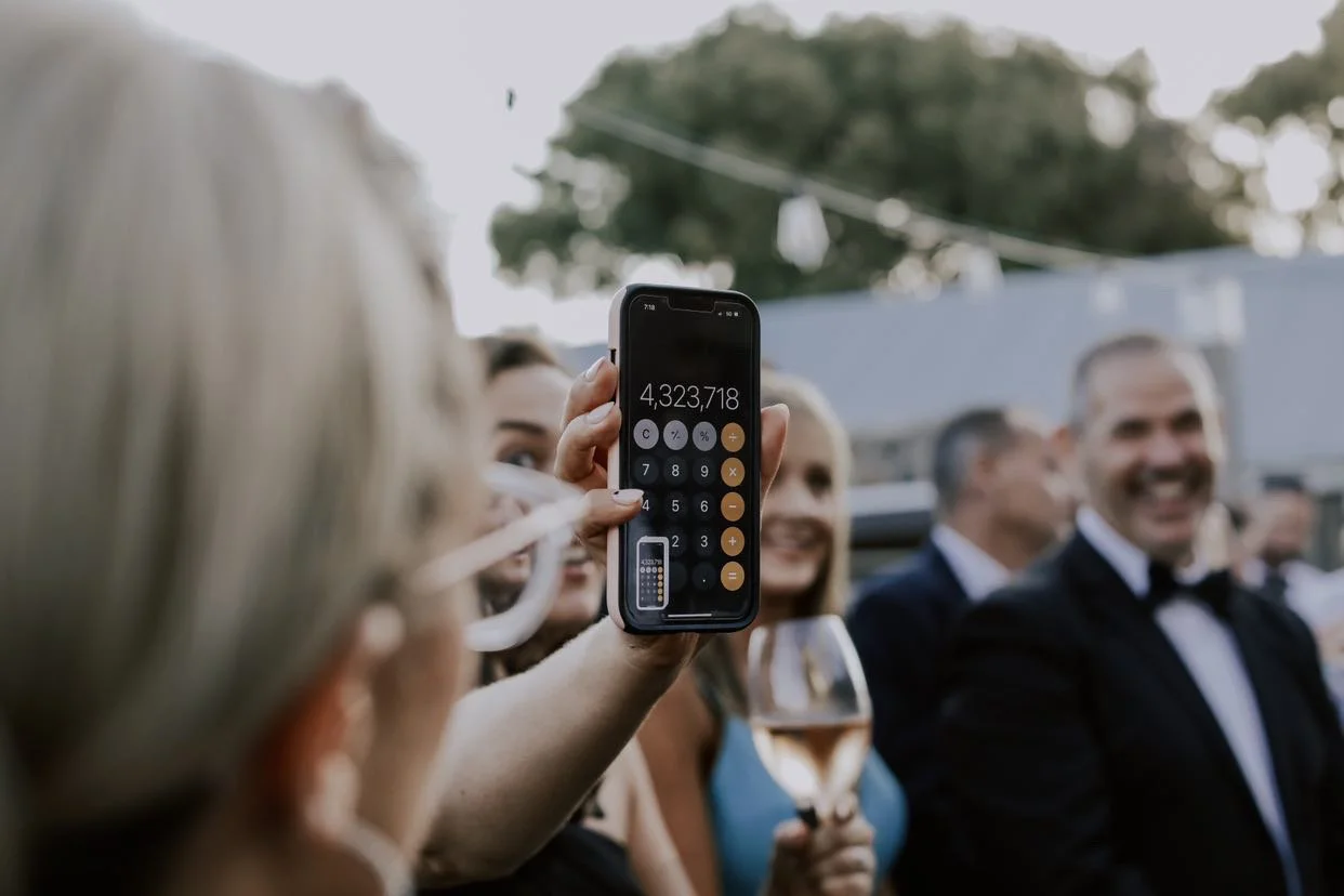 Close-up of a smartphone displaying 4,323,718 on a calculator app, held by a person among a group of people dressed formally at an outdoor event.
