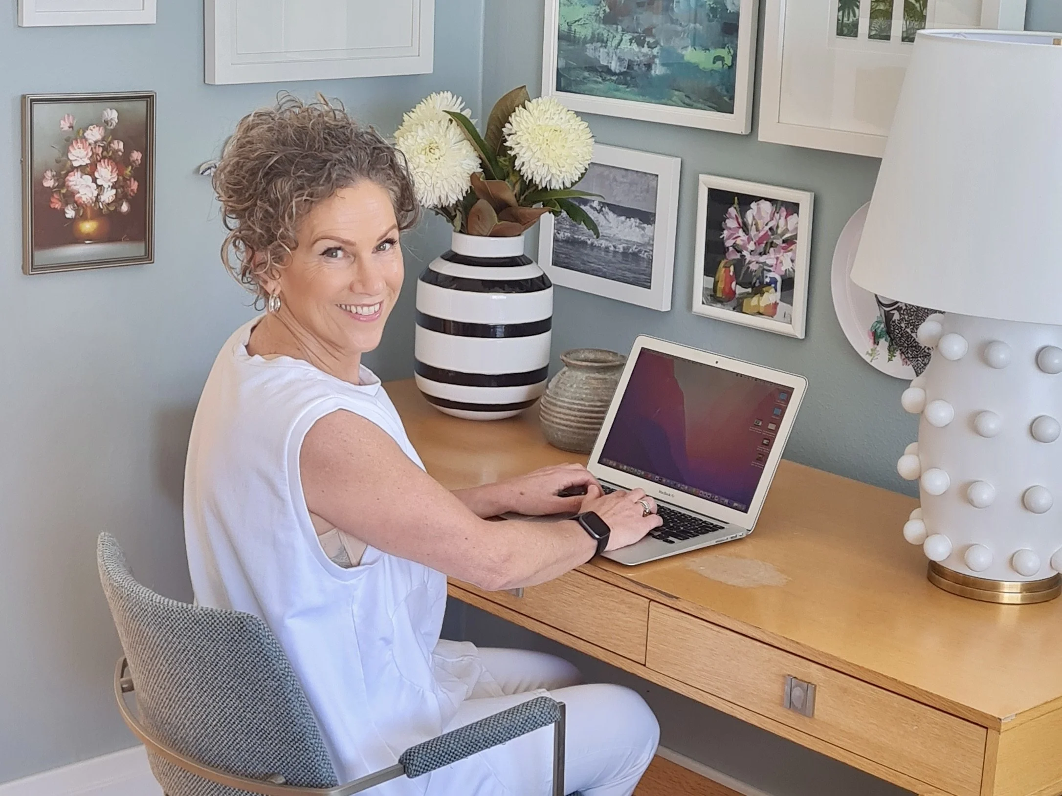 Caro with a curly ponytail smiling while sitting at a desk using a laptop in a decorated room with framed pictures and a large white lamp - Happy & Healthy with Caro