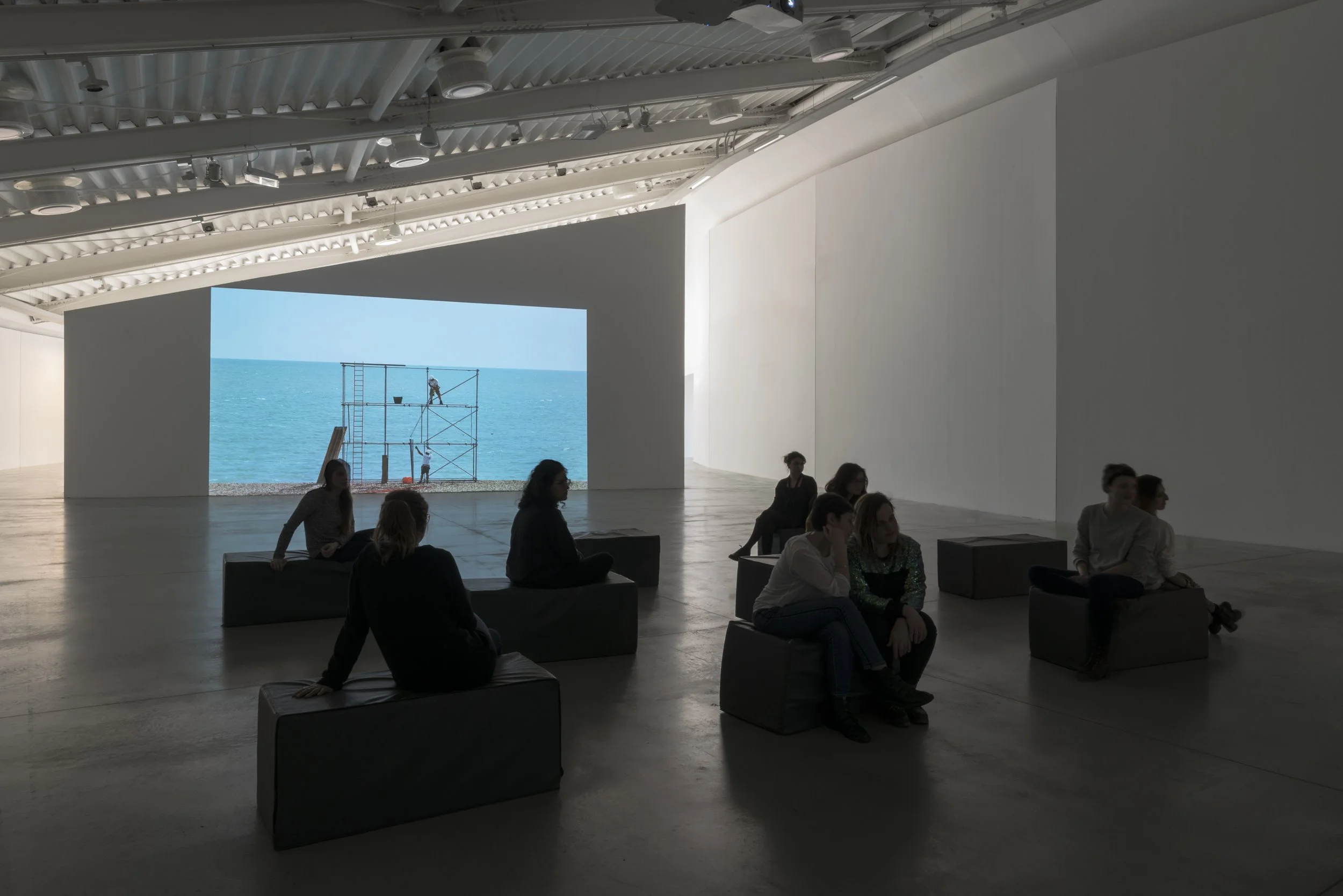 People seated on benches in an art gallery watching a large projection showing Mark Wallinger’s Construction Site, a single-channel colour video projection showing three professional scaffolders erecting and dismantling a structure on a beach.
