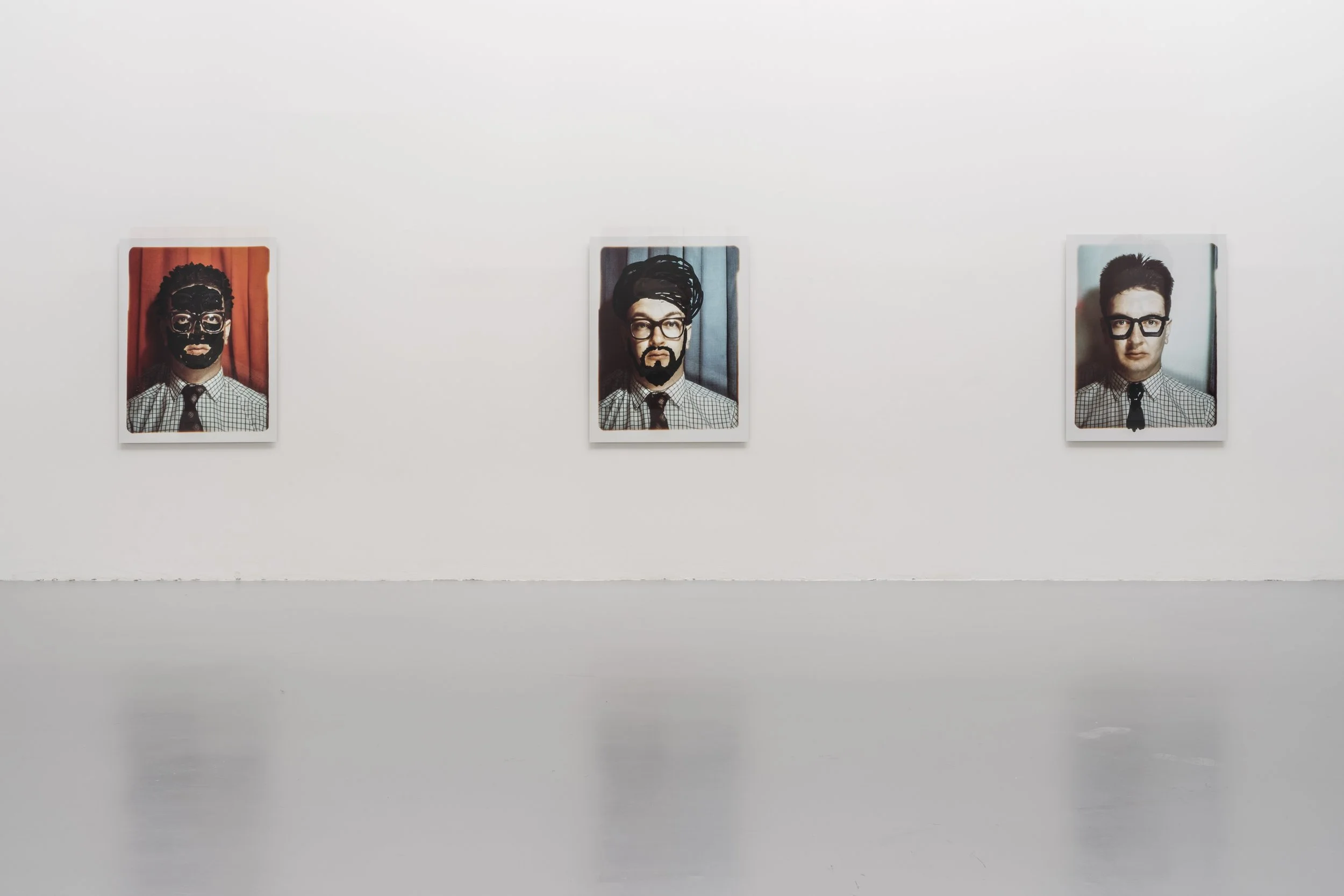 Three portraits of men in glasses hanging on a white gallery wall, with the middle man sporting a turban-like headwrap and a beard, and the other two men clean-shaven with different hairstyles.