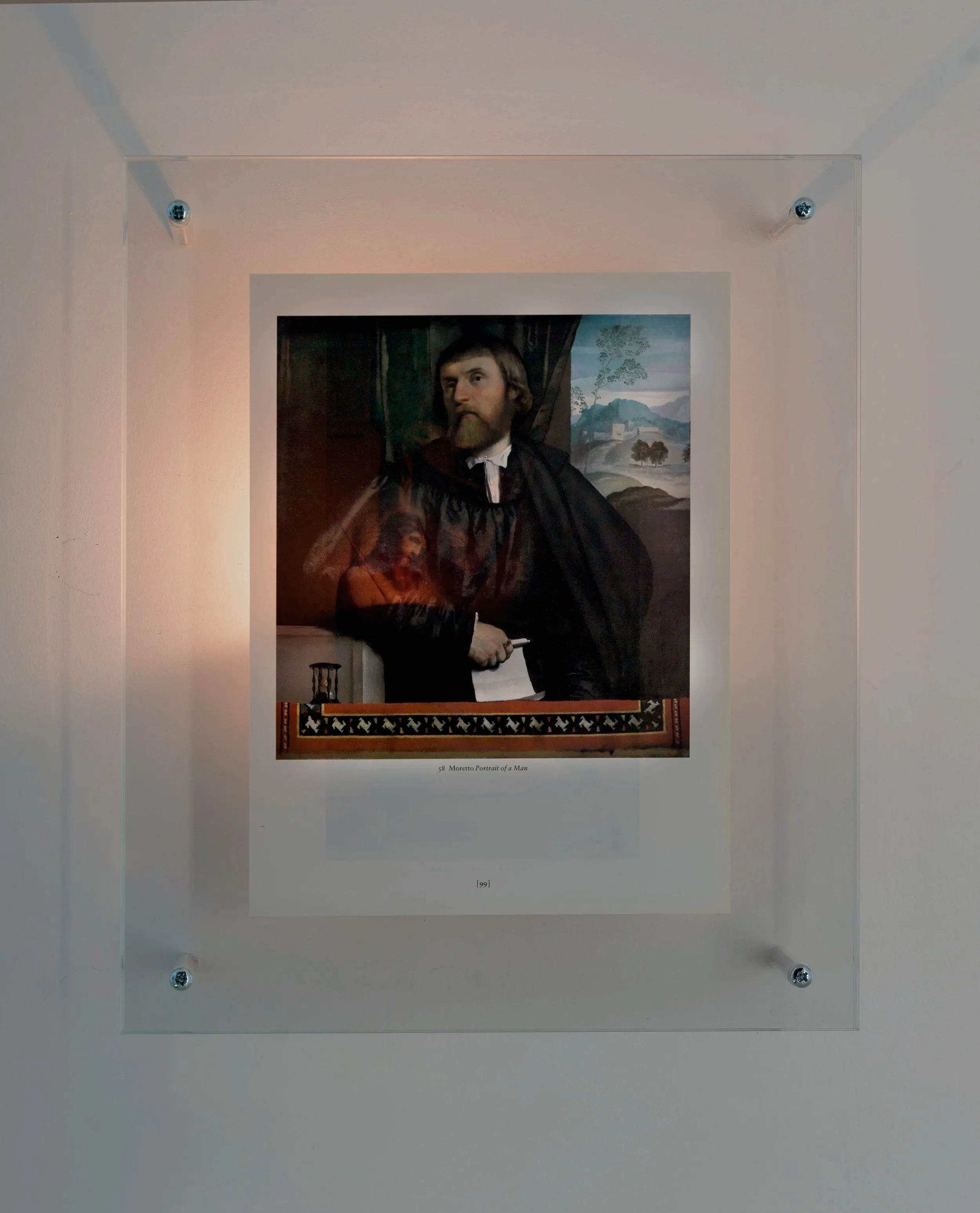 Detail of Mark Wallinger’s Genius of Venice (1991) framed art piece displayed on a gallery wall, sandwiched between glass and illuminated from behind by a flickering nightlight to reveal the presence of an image overleaf.