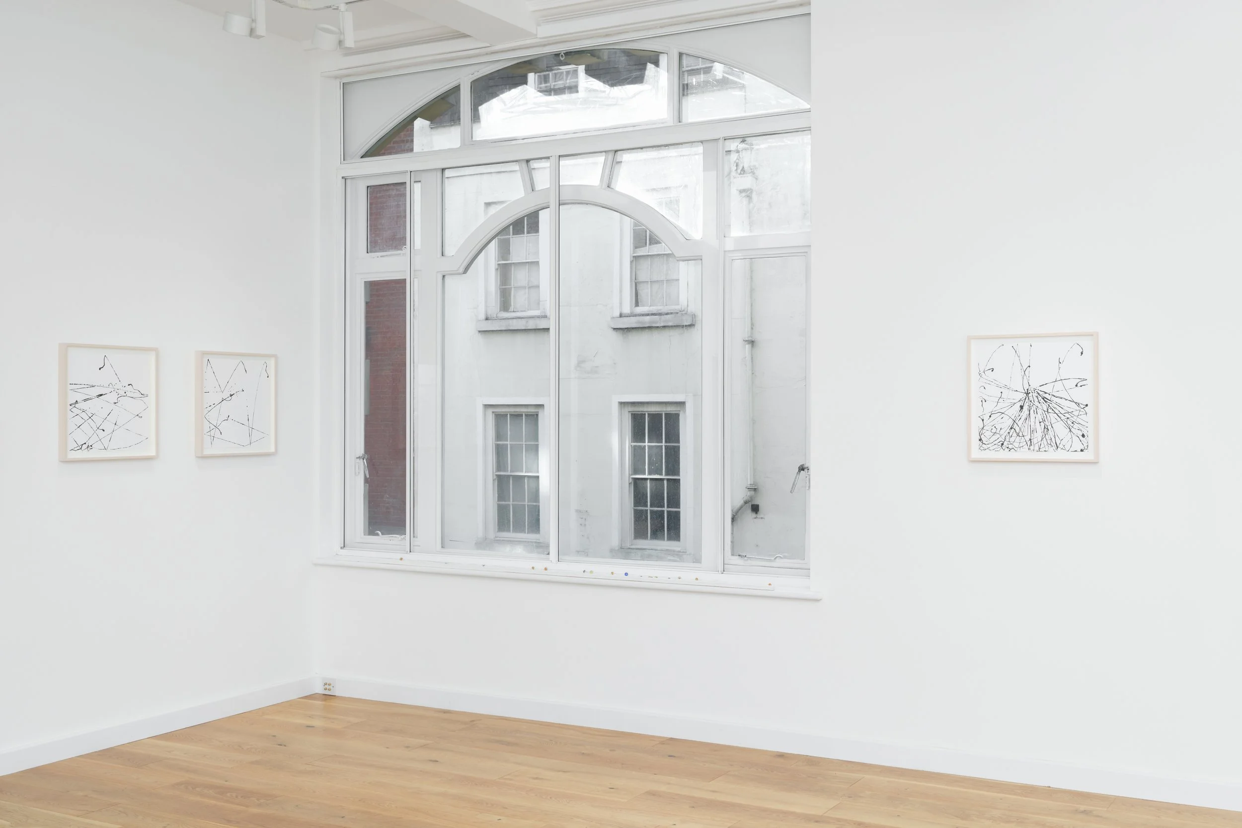 An art gallery with white walls and hardwood floors, featuring three framed abstract line drawings on the walls and a large window with archedtop and multiple smaller panes, showing a brick and white building exterior. Mark Wallinger’s at Cedric B