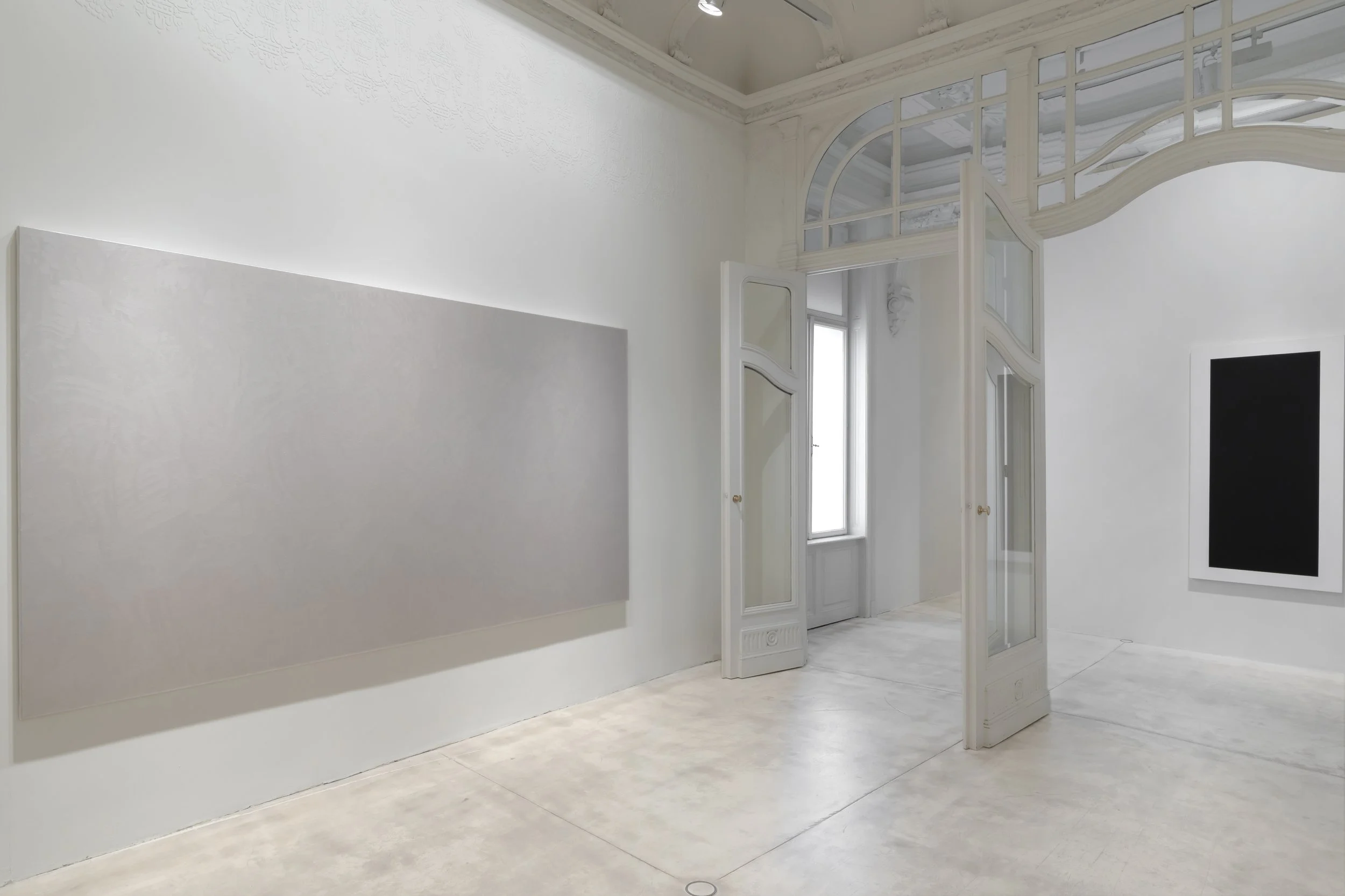 Interior of an art gallery with white walls, a blank light-colored painting, a black frame on the wall, and a decorative archway with glass panels.