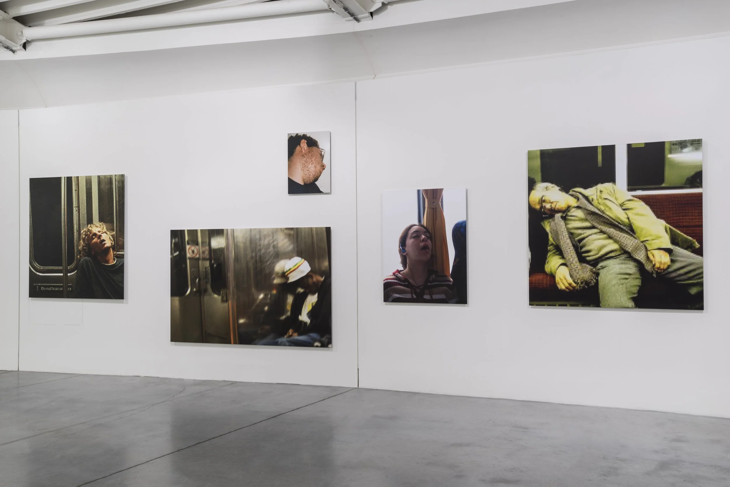 Art Gallery featuring Mark Wallinger’s "The Unconscious", enlarged images sourced from the Internet of people sleeping on public transit, displayed on a white wall in an art gallery.
