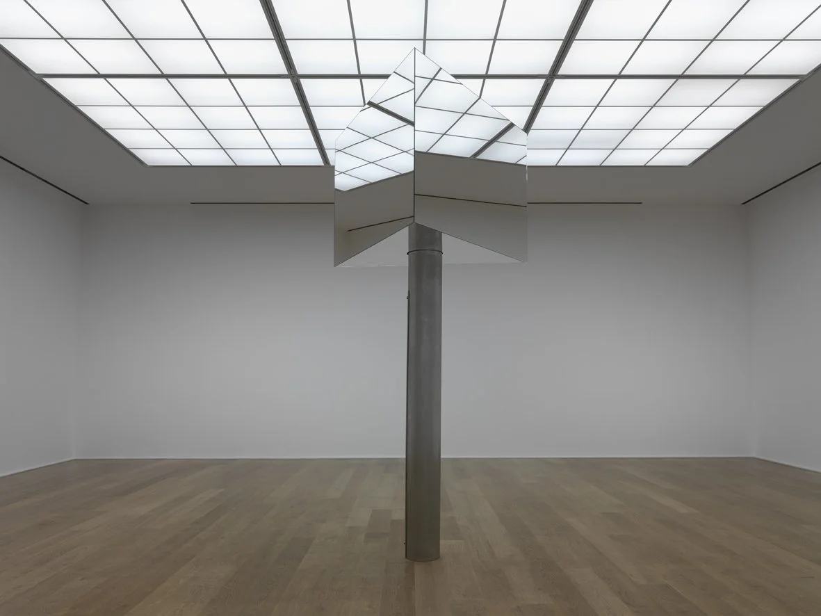Inside a white gallery with a grid-patterned ceiling, Mark Wallinger’s Superego (2016) stands alone. This mirrored sculpture, inspired by the revolving New Scotland Yard sign, evokes authority and vigilance, reflecting viewers while remaining distant
