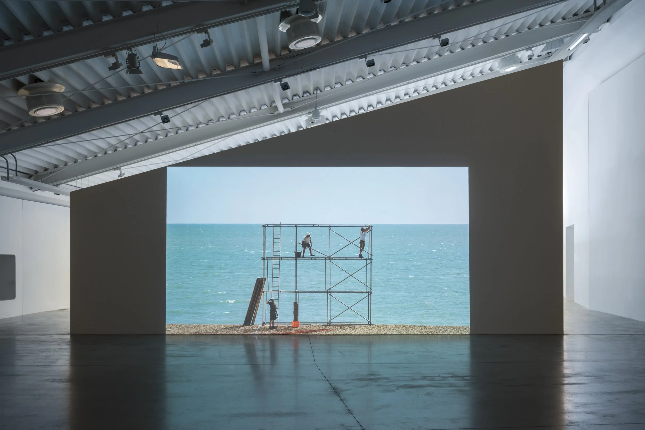 View of a large projection showing Mark Wallinger’s Construction Site, a a single-channel colour video projection showing three professional scaffolders erecting and dismantling a structure on a beach, against a backdrop of sky, sea and land.
