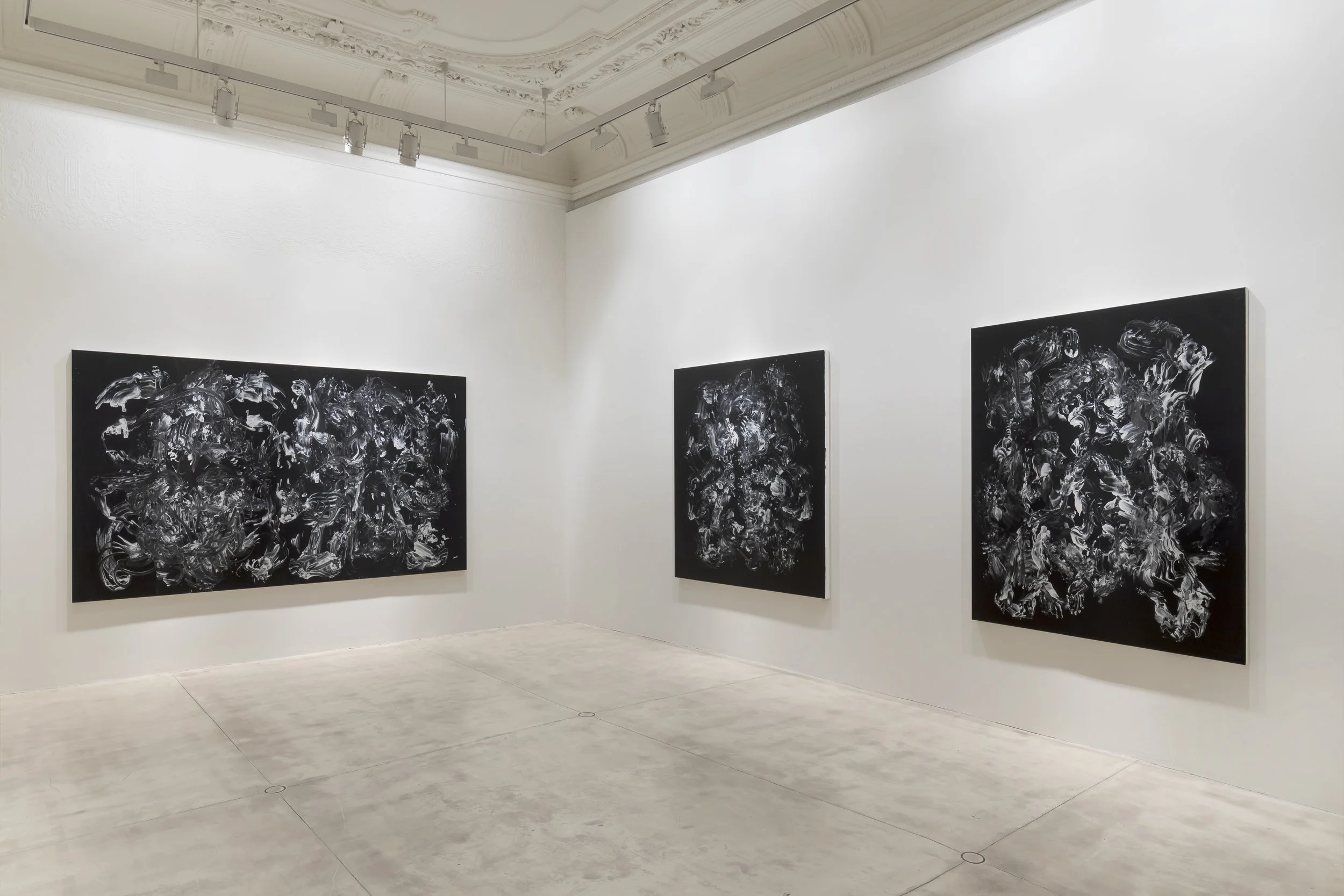 Three large abstract black and white paintings displayed on a white wall in a gallery, with minimal lighting and high ceiling.