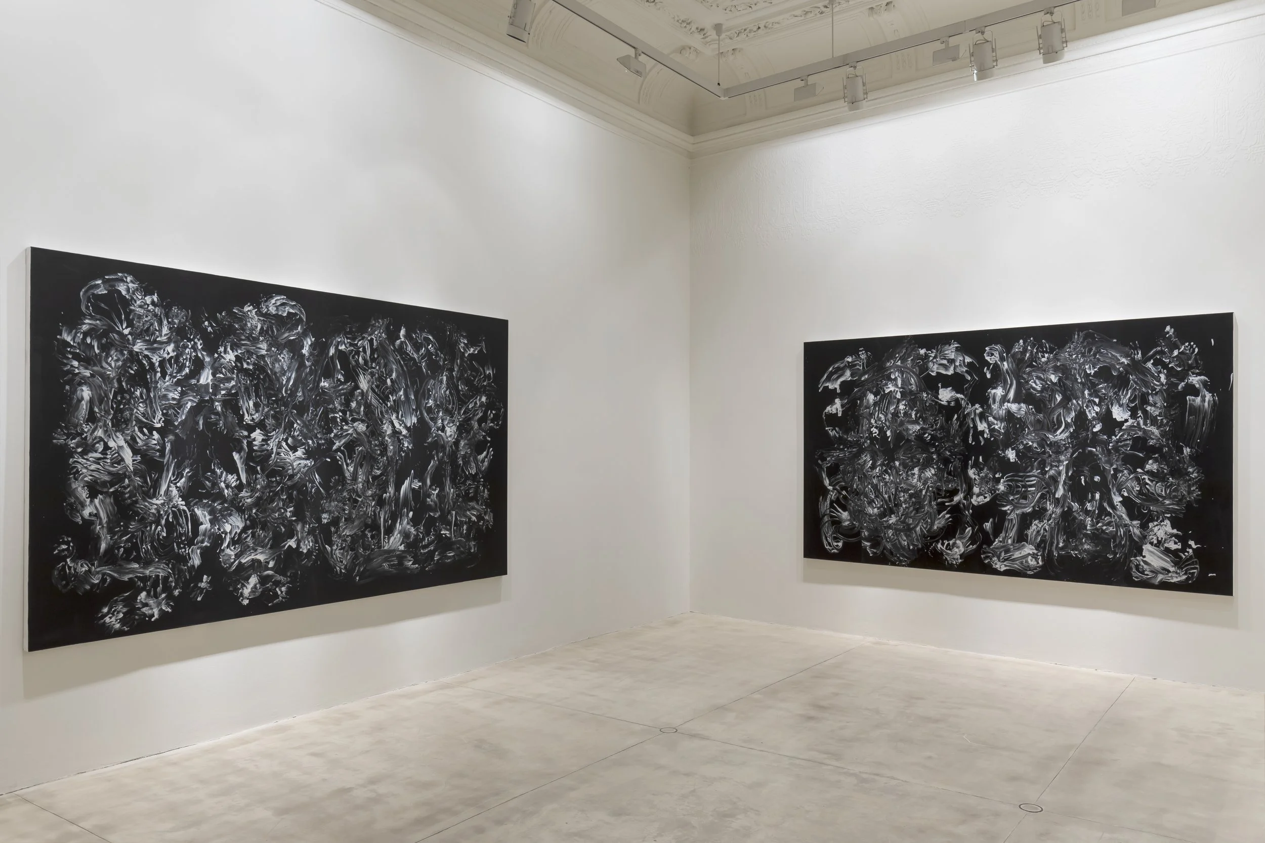 Two large black and white abstract paintings hanging on white gallery walls in an art gallery, with a light-colored floor and track lighting overhead.