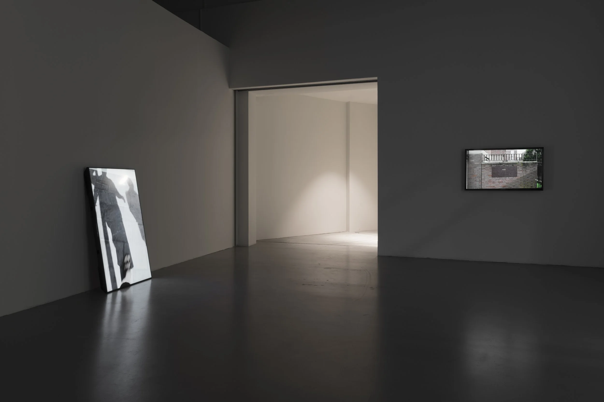 An art gallery featuring dark flooring and white walls showcases Mark Wallinger's Shadow Walker and MARK on screens, each mounted on separate walls, with a doorway leading into a brightly lit room.