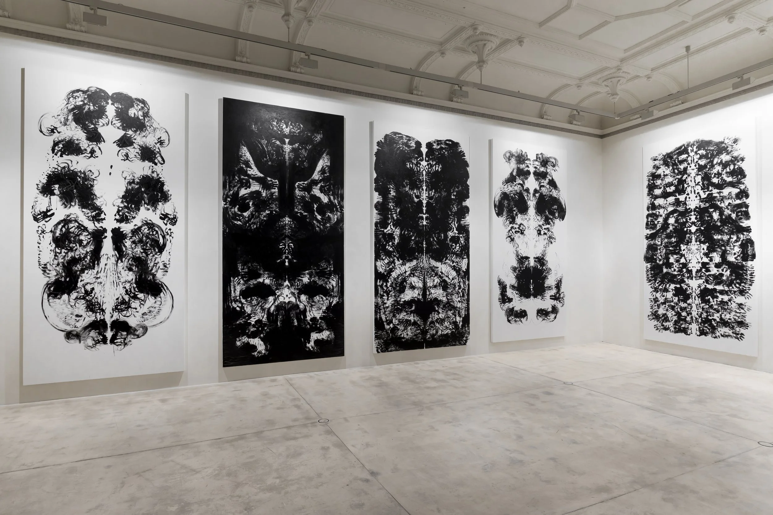 Five large black and white abstract paintings on display in an art gallery with white walls and a concrete floor.