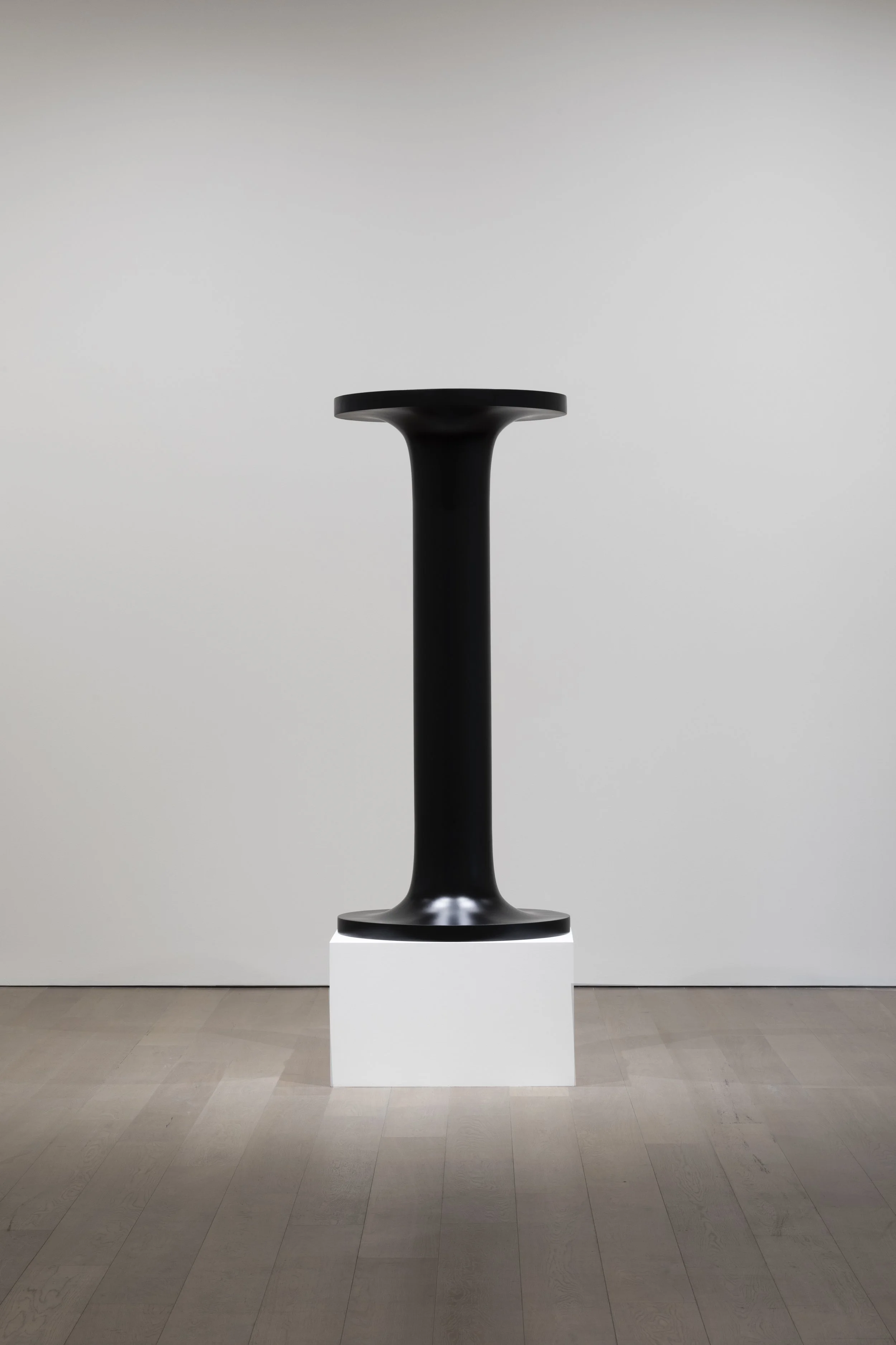 A black modern pedestal sculpture on a white block, displayed in a gallery setting with wooden flooring and white walls.