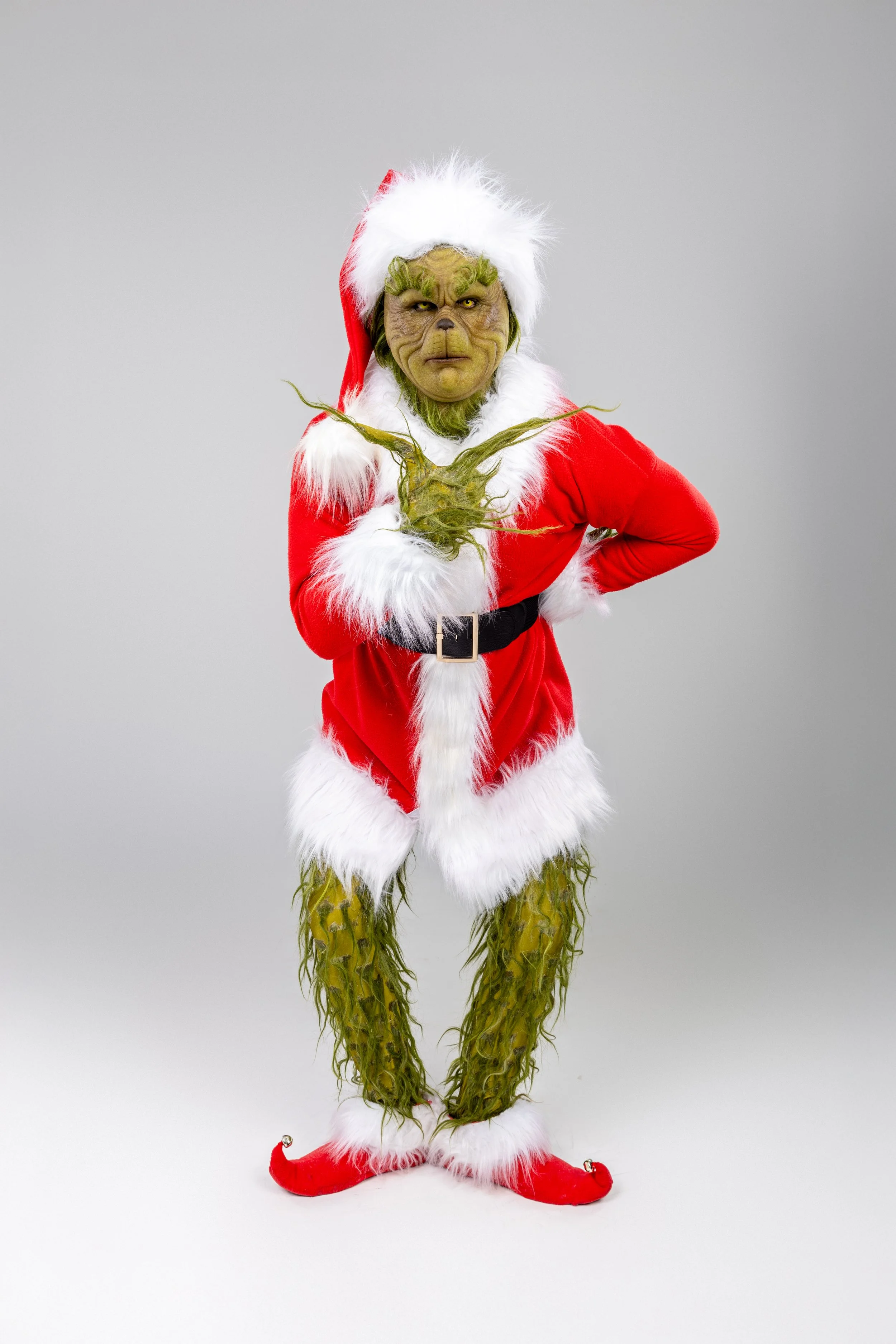 A person dressed as the Grinch in a Santa outfit, with green fur, standing against a plain gray background.
