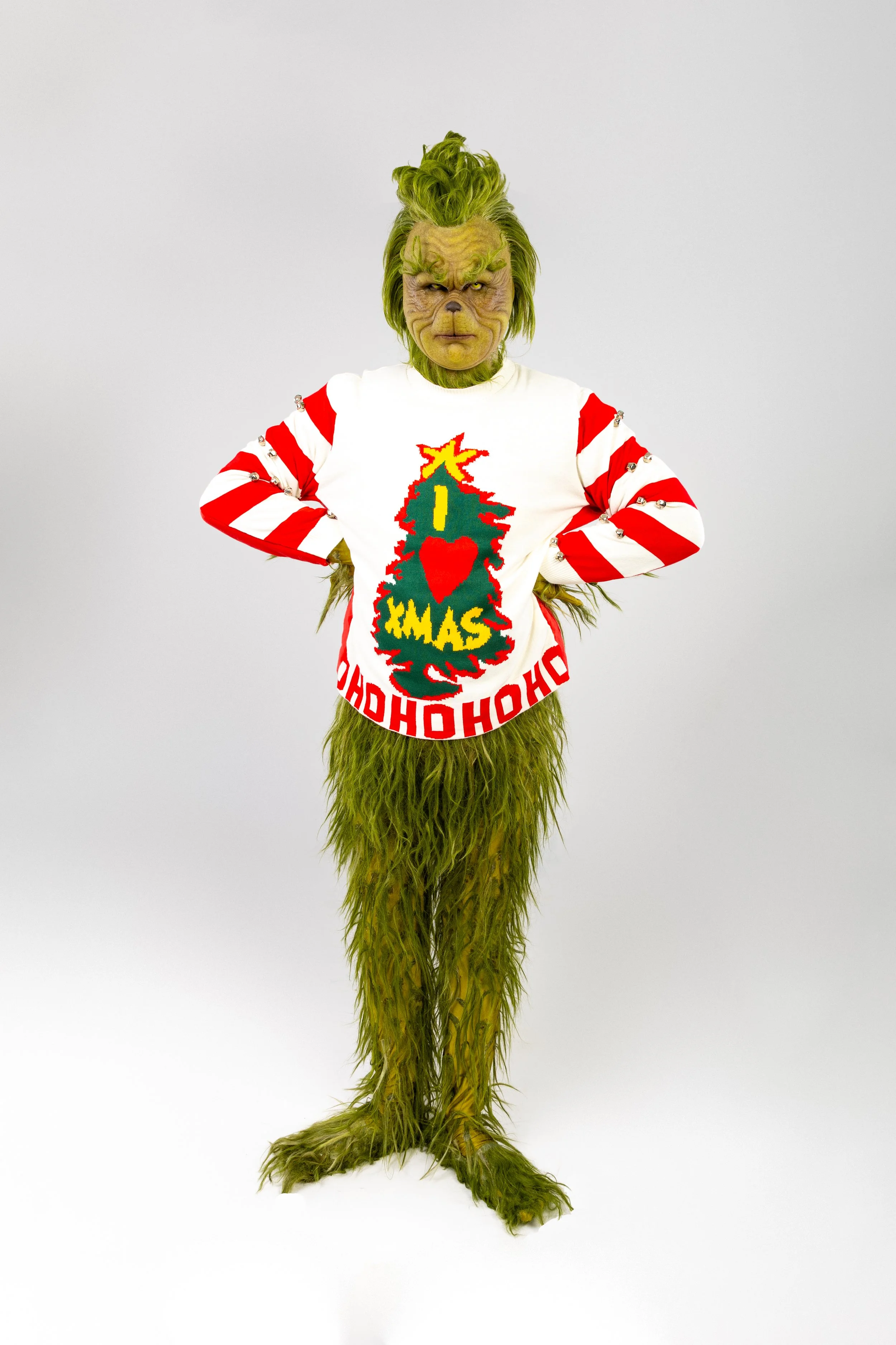 A person dressed in a Grinch costume with a green furry body and a mask, wearing a Christmas sweater and standing with hands on hips against a plain background.