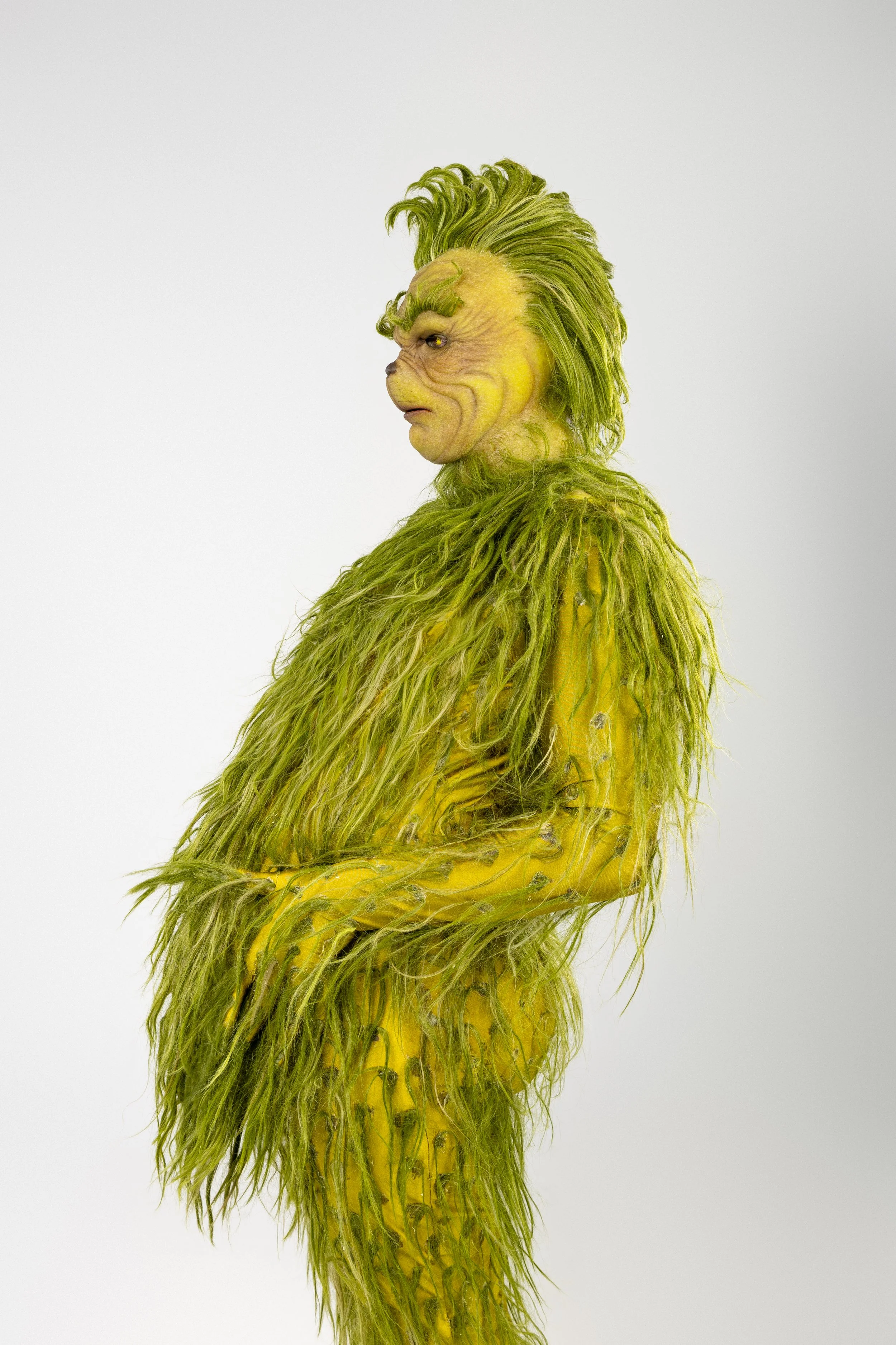 A person dressed in a full-body costume resembling the Grinch, with green fur, a wrinkled face, and wild hair, standing in profile against a plain white background.