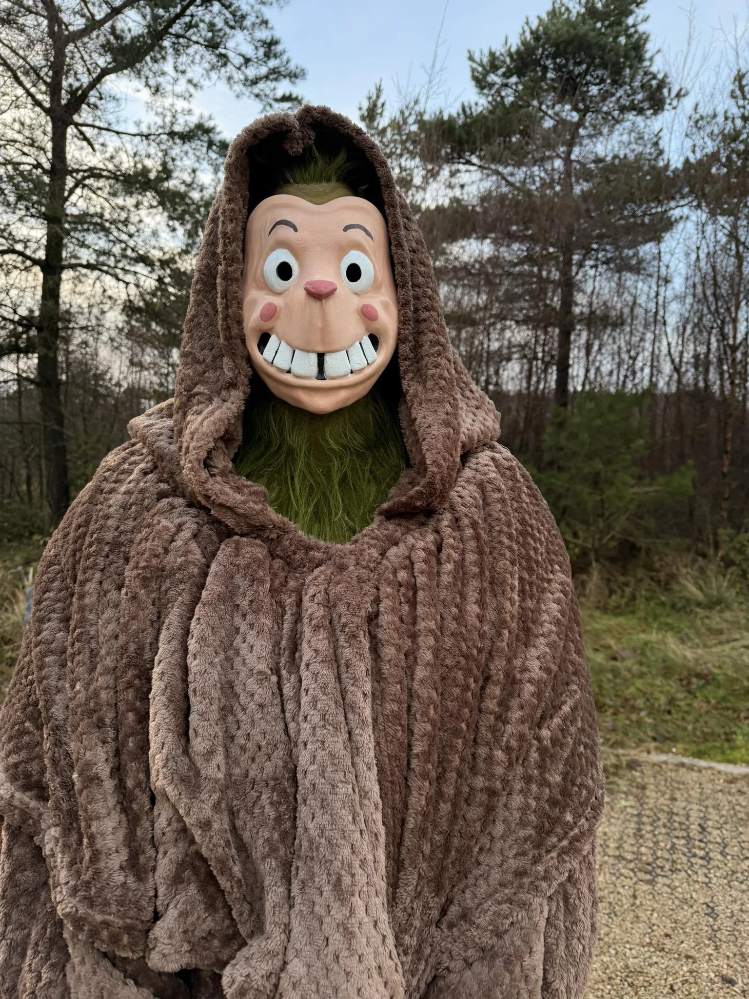 Person wearing a costume with a large exaggerated smiling mask and a brown, furry cloak, standing outdoors with trees and a cloudy sky in the background.
