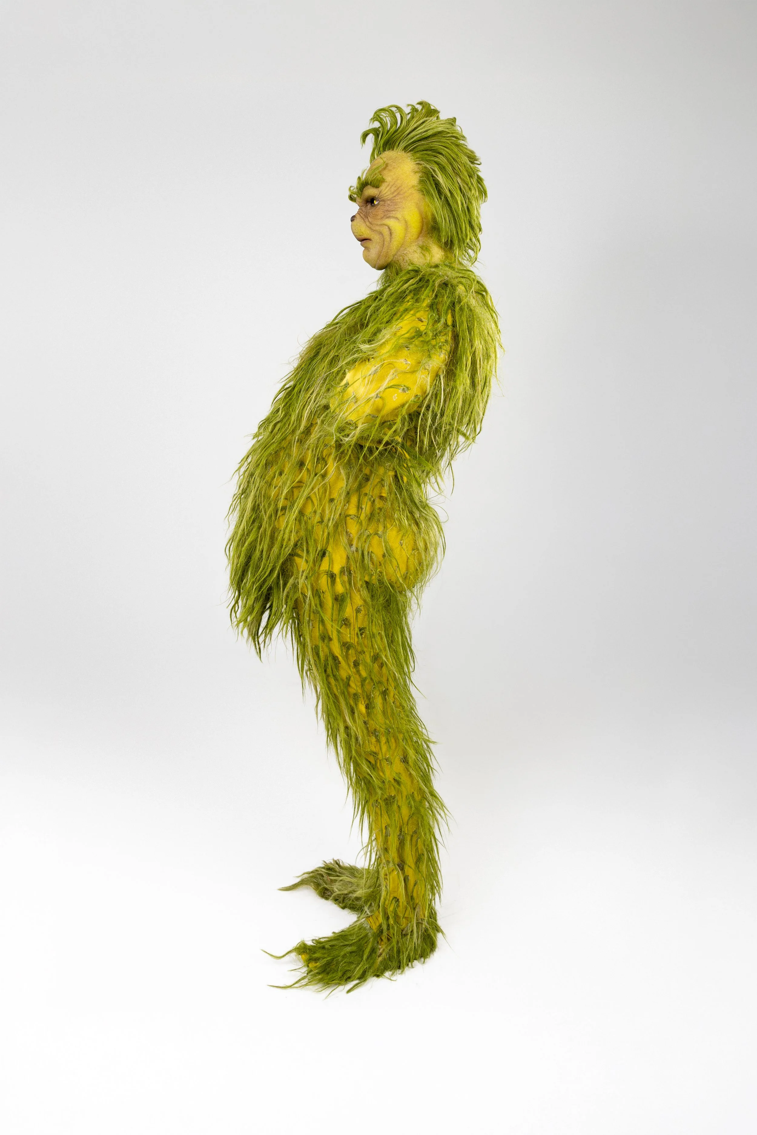 Person in a detailed costume resembling the character from 'The Grinch', covered in green fur and yellow accents, standing sideways against a plain white background.