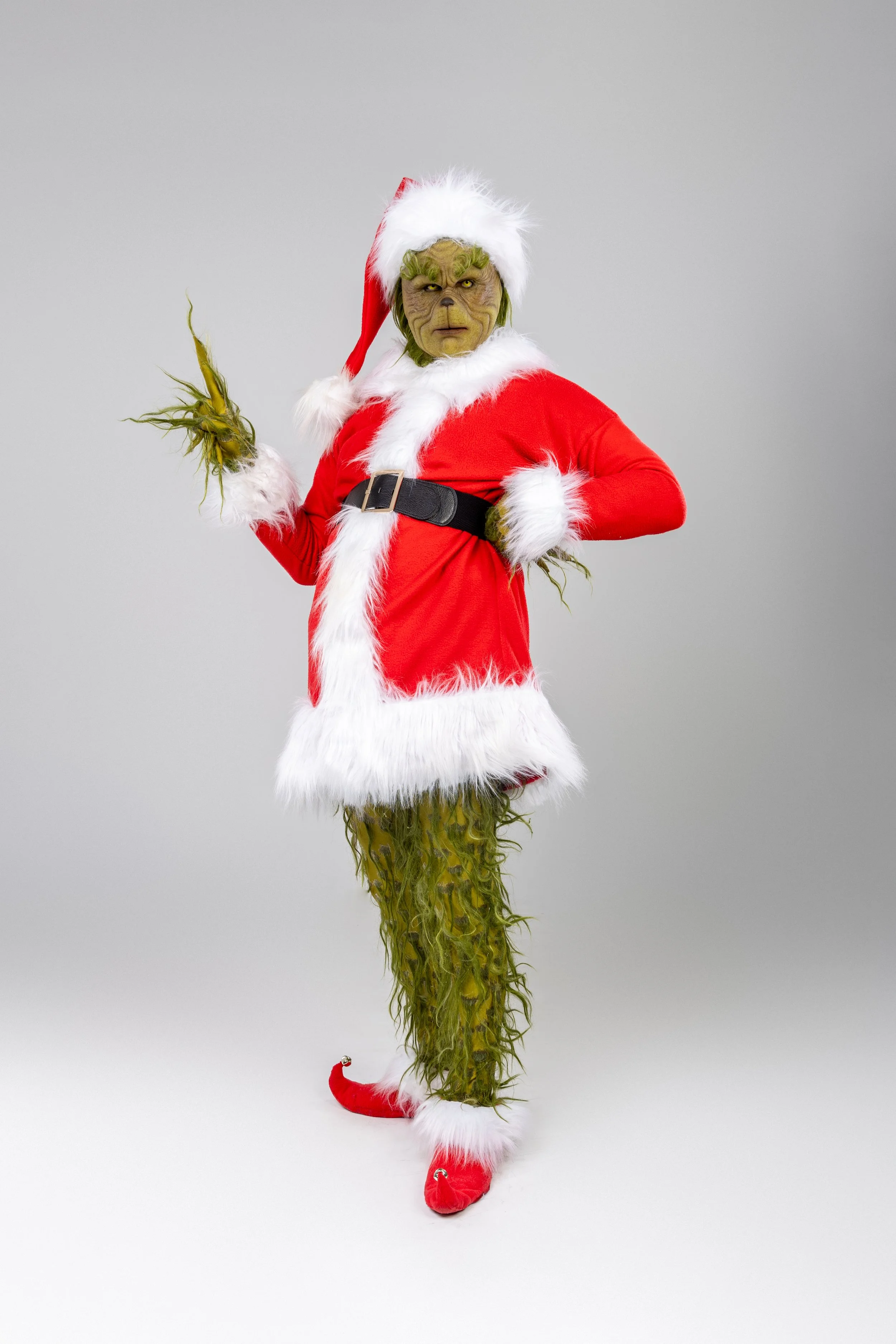The Grinch dressed in a festive Santa costume with red suit, white fur trim, Santa hat, and red shoes, standing against a plain gray background.