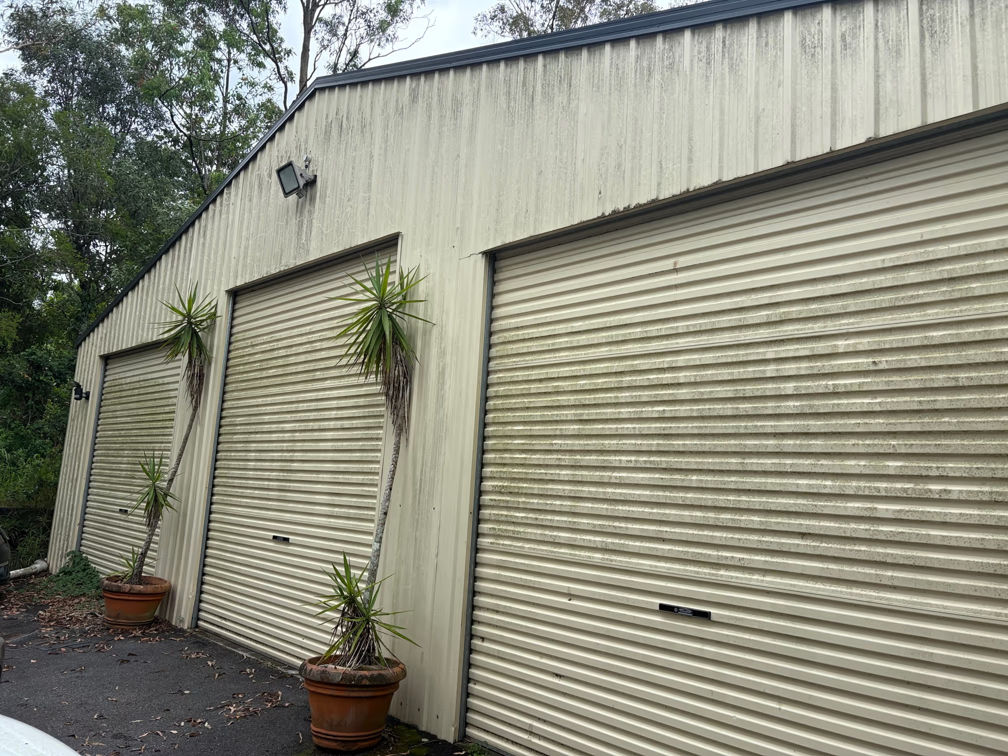 The image shows a beige industrial building with two roller shutters, some potted plants in front, and a security camera mounted on the upper left side. The building appears weathered with visible dirt and staining.