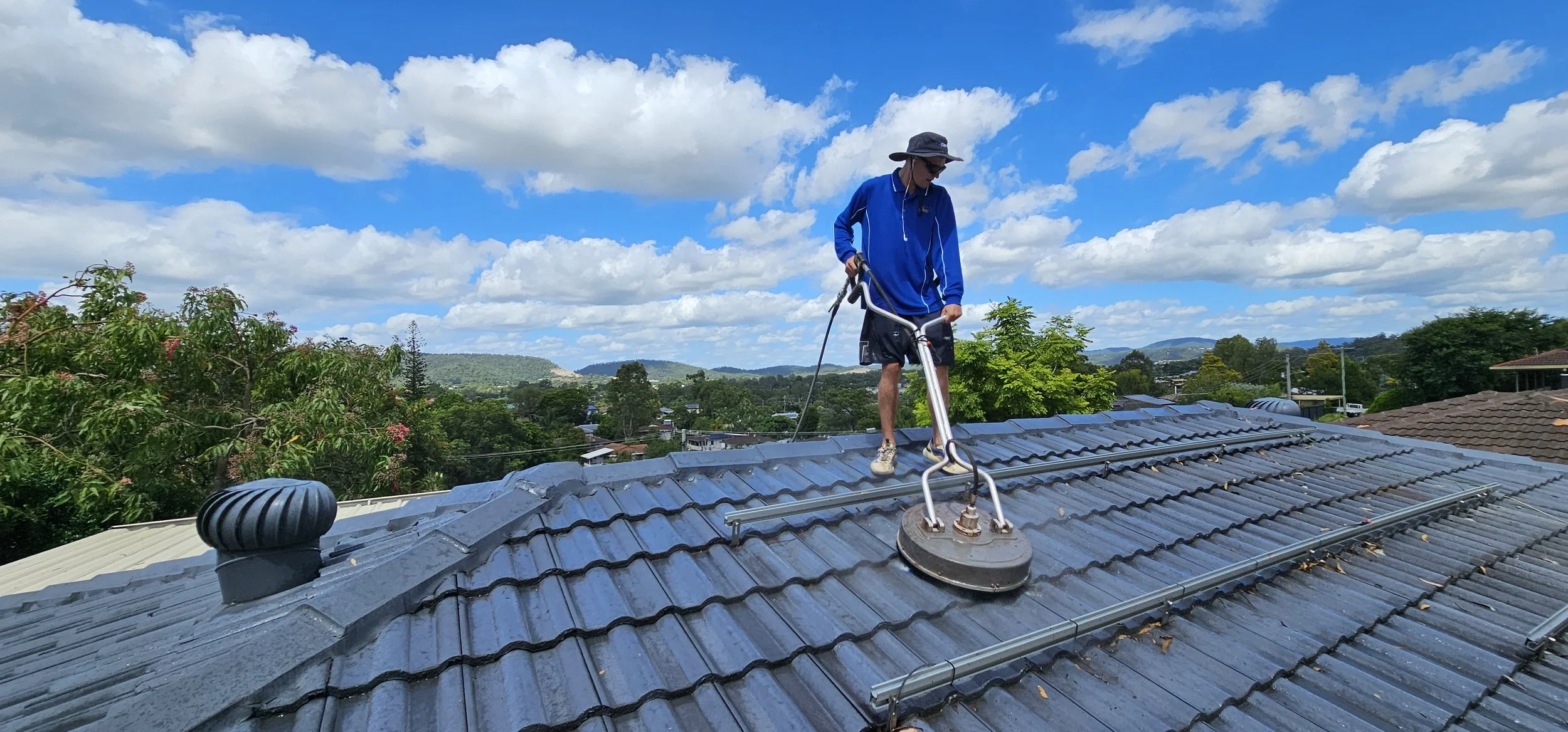 Person cleaning a rooftop with a pressure washer, wearing a blue shirt and hat, surrounded by trees and a cloudy sky.