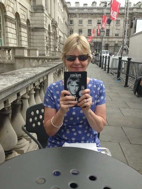 A woman with blonde hair wearing sunglasses and a blue polka dot shirt, taking a selfie with a book in an outdoor public space, with historic buildings and red flags in the background.