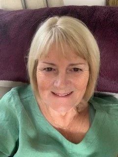 A smiling middle-aged woman with blonde hair, wearing a light green top, sitting on a purple couch.