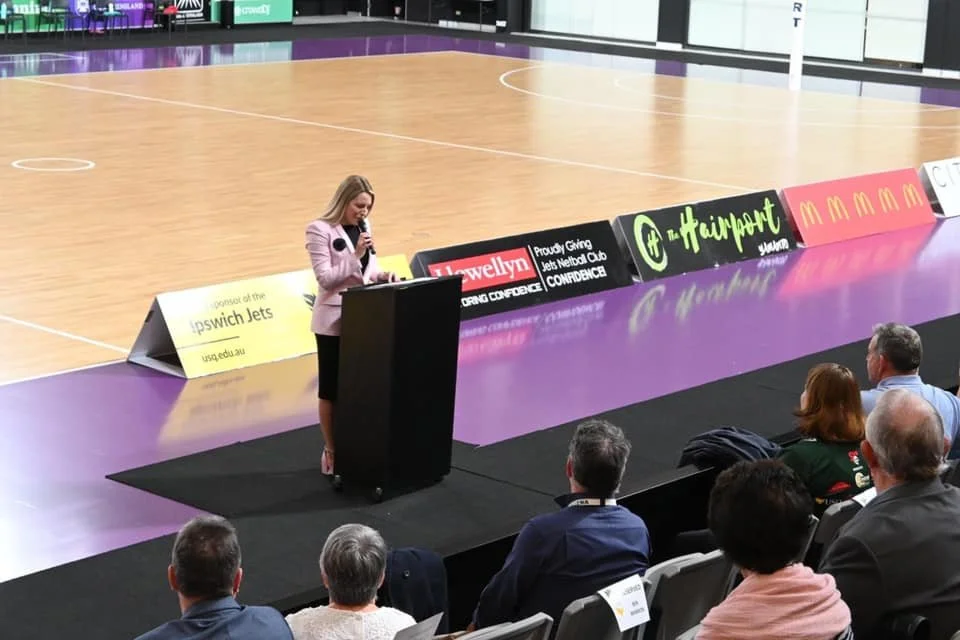 A woman in a pink blazer speaking at a podium during a conference in an indoor sports arena, with audience members seated in front and advertising banners displayed along the side of the court.